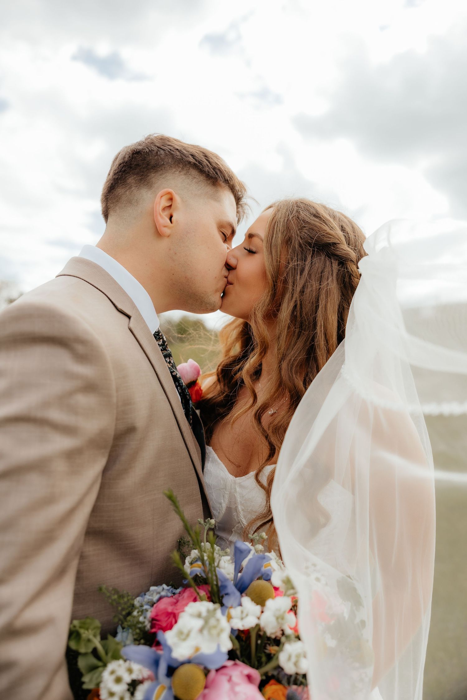 Bride and groom share a romantic kiss outdoors, with bridal veil flowing and colorful bouquet, on their wedding day.