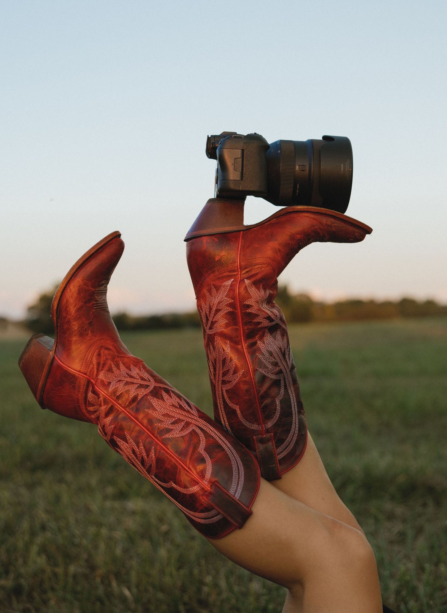 Red leather cowboy boots with a camera lens balanced between them in a grassy field.