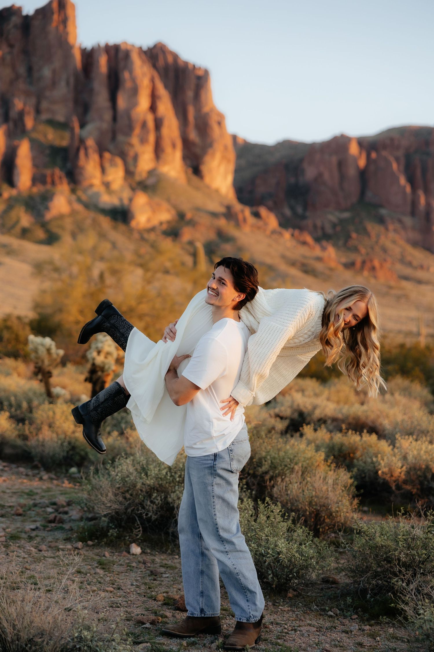 A couple embraces playfully against a backdrop of dramatic red rock formations in the desert during golden hour.