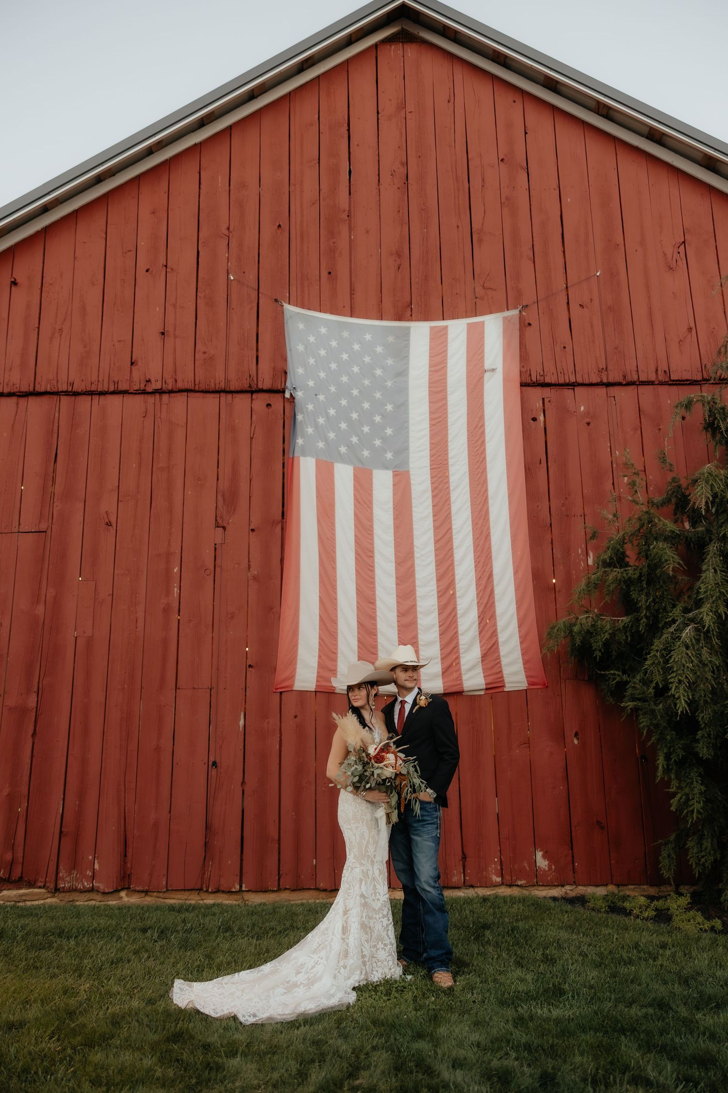 Bride and groom in cowboy hats pose before a red barn with an American flag at a rustic country wedding.