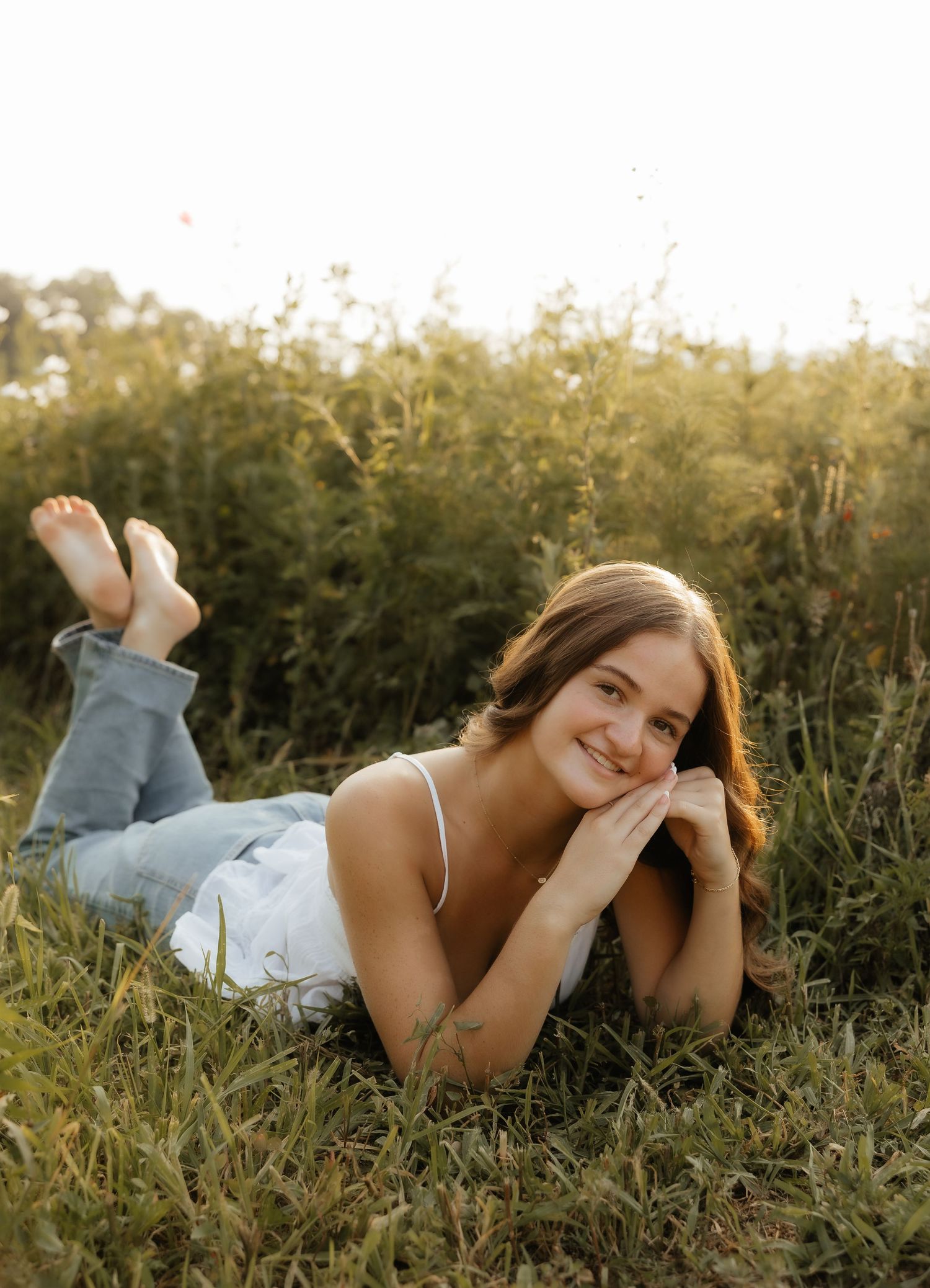 Young woman in white top and jeans lying in a sunlit meadow, smiling warmly at the camera.