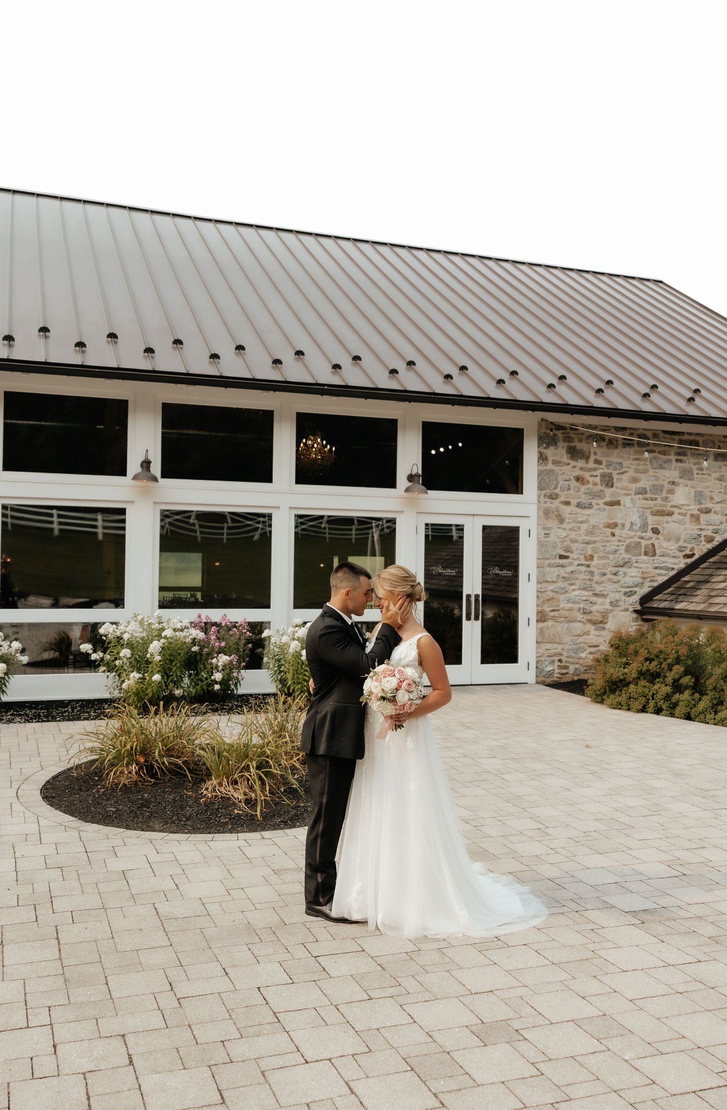 Bride and groom share a romantic moment outside a modern stone wedding venue with large windows.