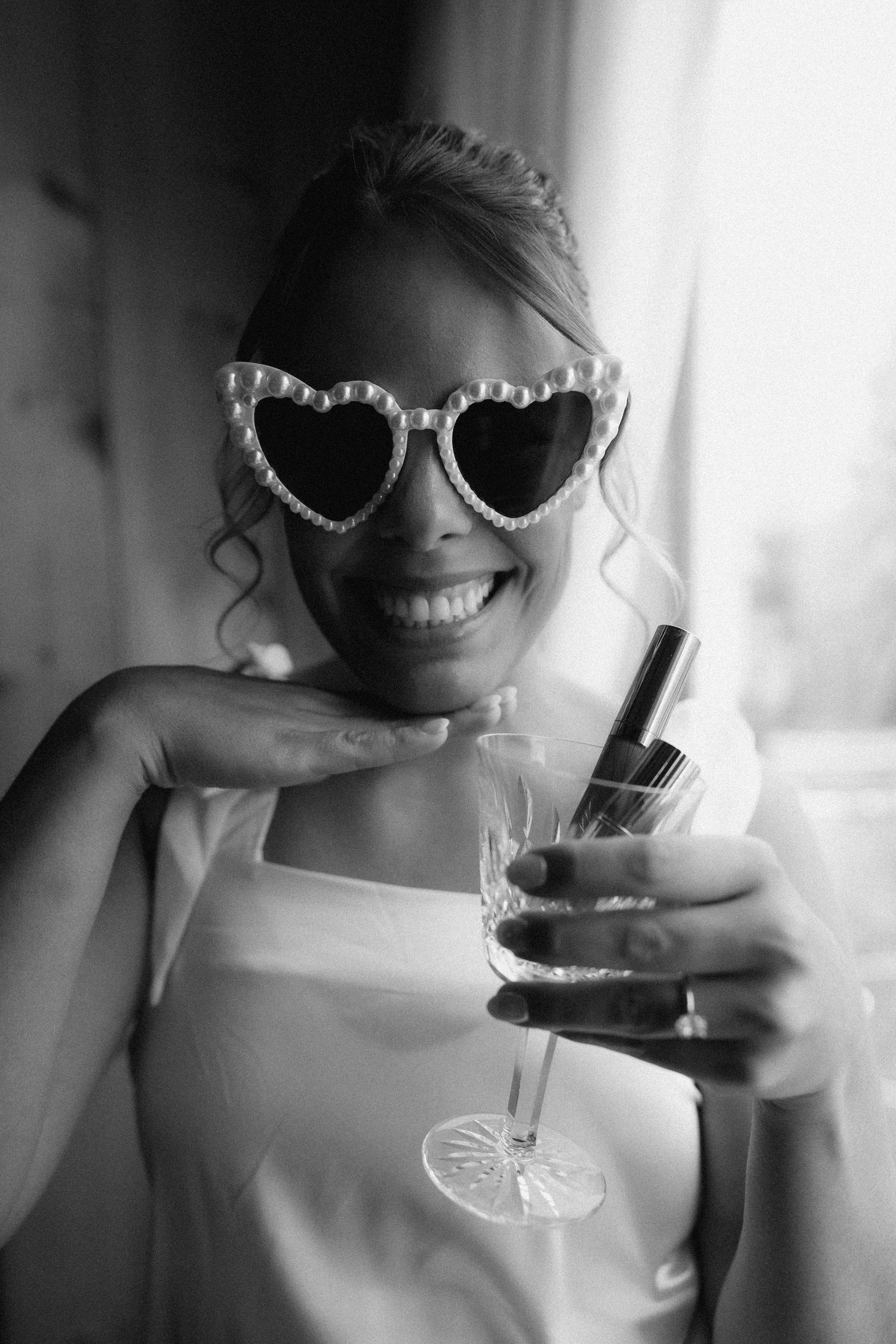 Smiling bride in heart-shaped pearl sunglasses holding a champagne glass, black and white wedding photo.