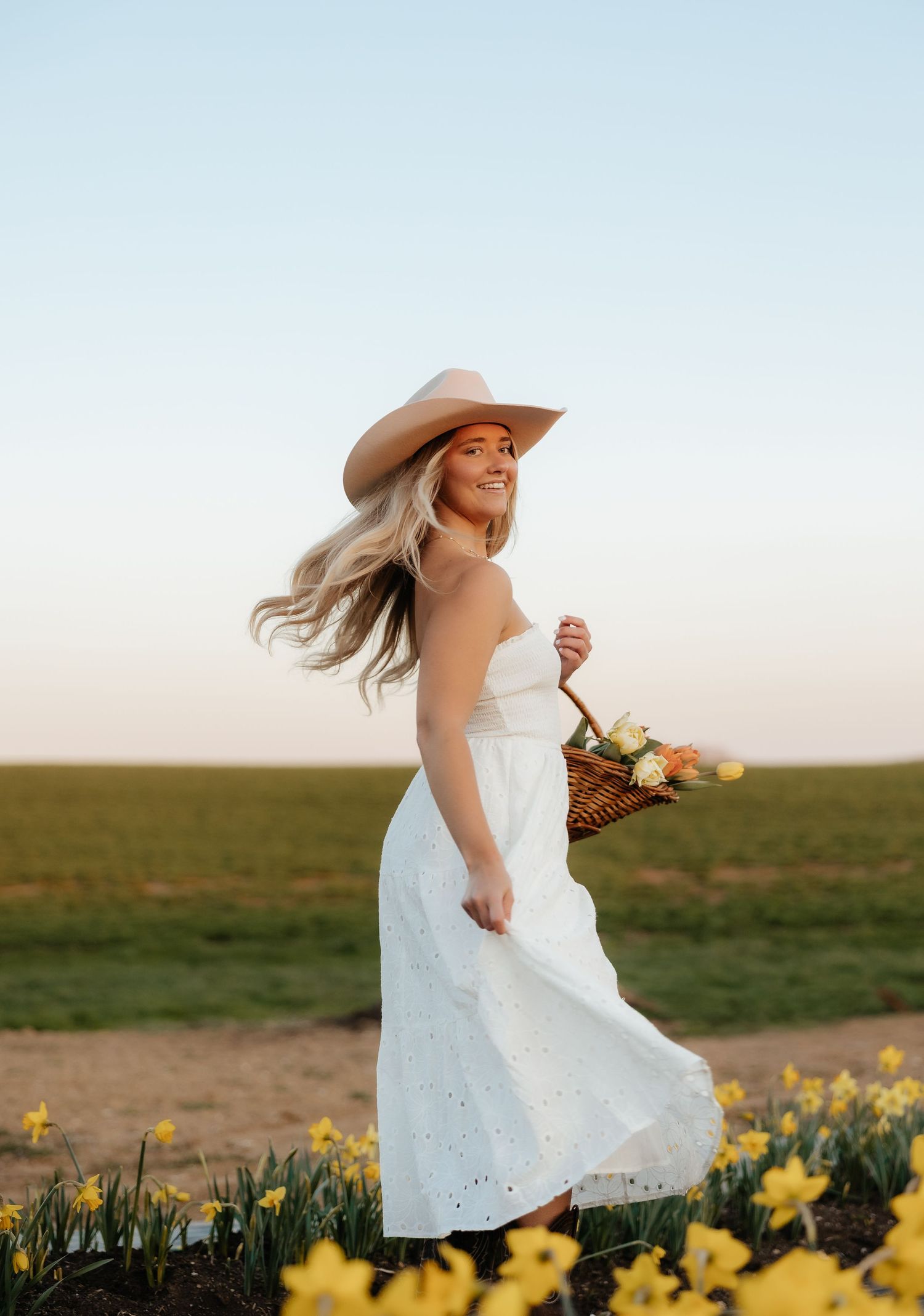 Woman in white sundress and wide-brim hat holding flower basket among yellow daffodils in a sunny field.