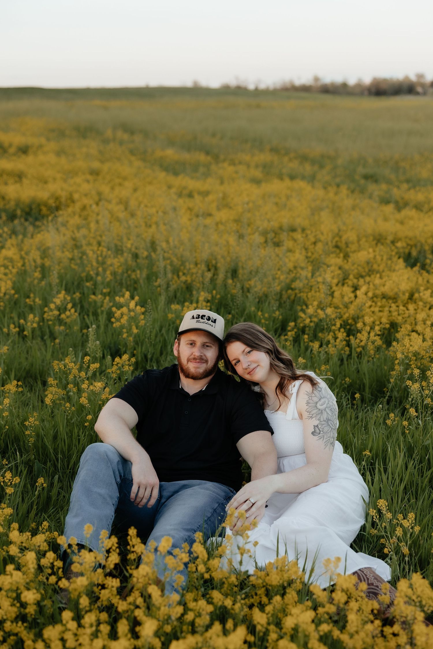 Couple sitting together in a vibrant yellow wildflower field, woman in white dress leaning on man in black shirt and cap.