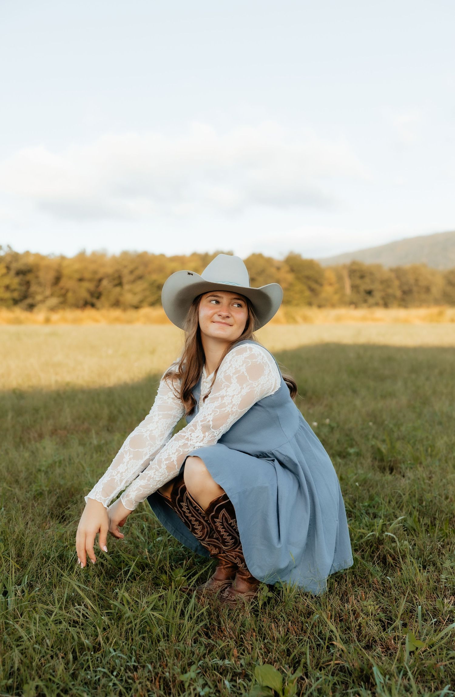 Young woman in cowboy hat, lace top, blue skirt, and brown boots crouching in a green field with trees behind her.