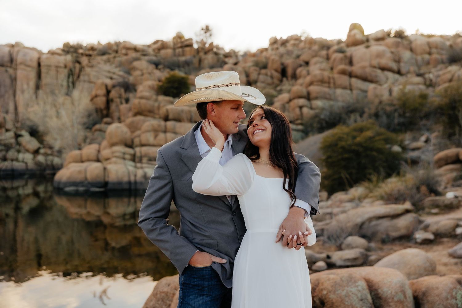 A couple in western attire embraces near a scenic desert lake surrounded by large granite boulders at sunset.