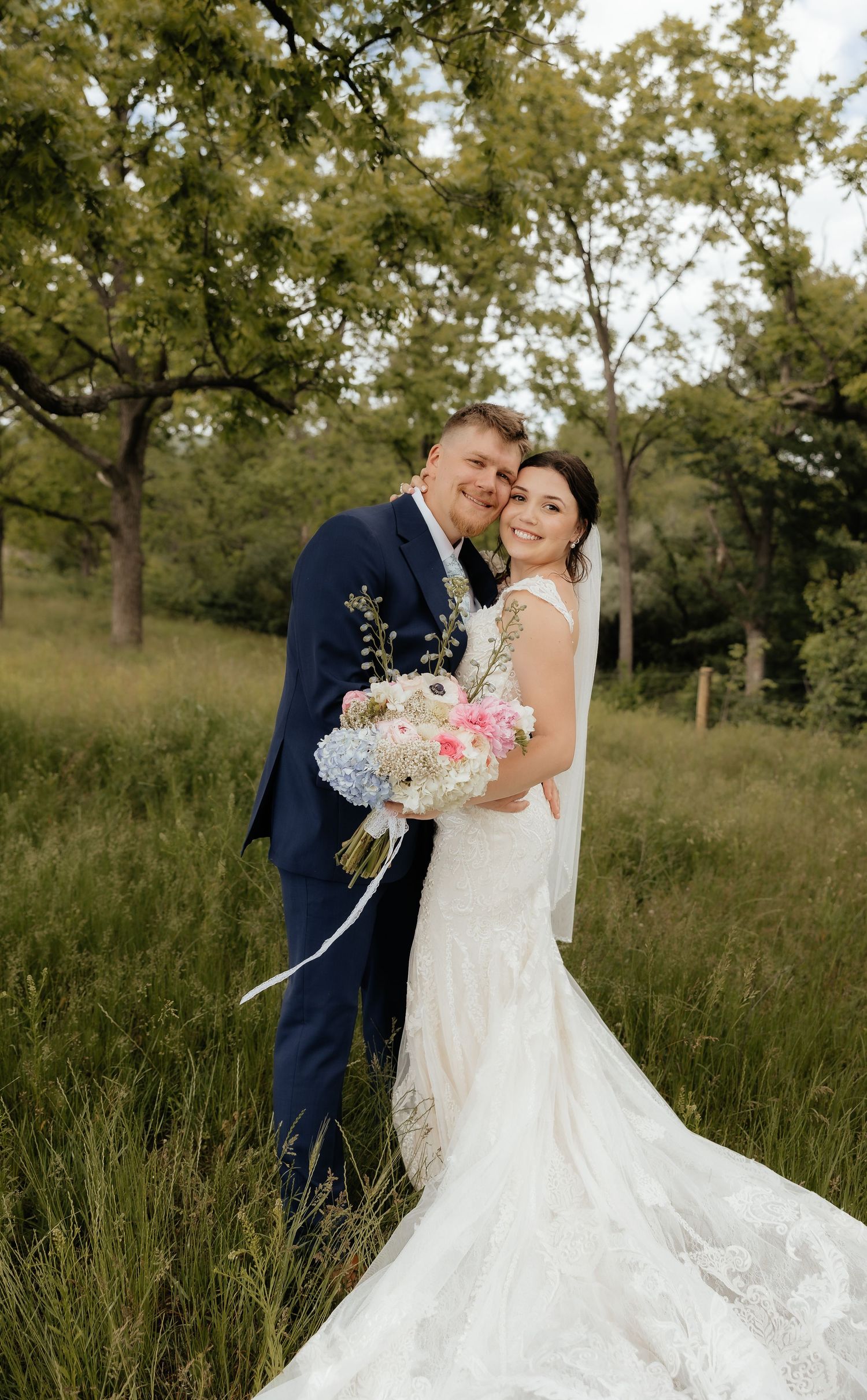 Bride in lace gown and groom in navy suit embrace outdoors holding pink and blue floral bouquet.
