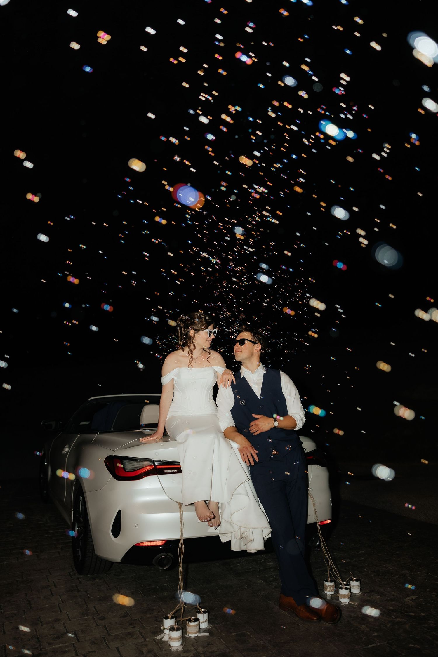 Bride and groom sitting on white car at night with colorful confetti falling around them.