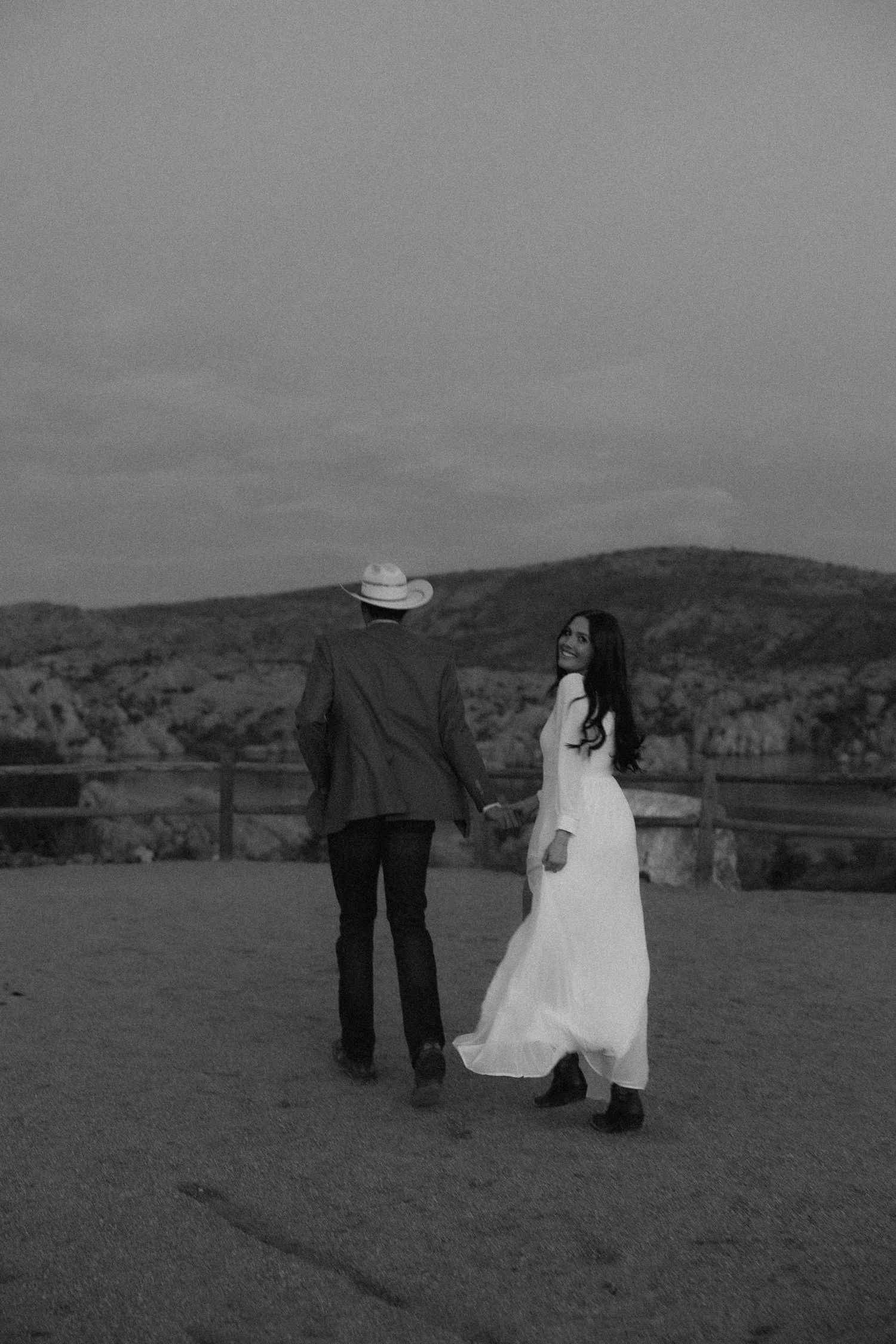 A black and white photo of a couple in western attire standing on a hilltop overlooking mountains.