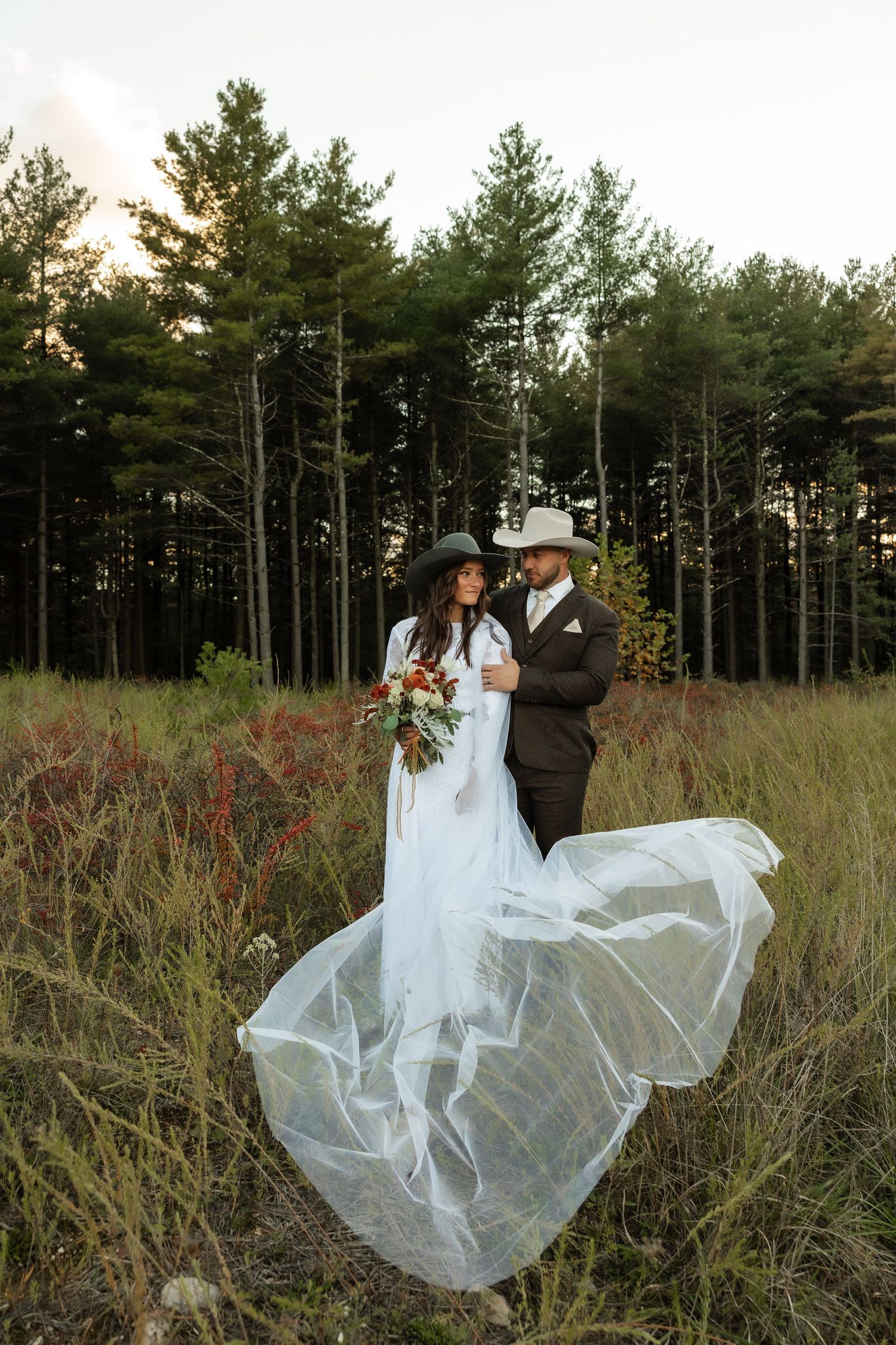 A couple in wedding attire stands in a field with tall pine trees at sunset as the bride's dress flows in the wind.