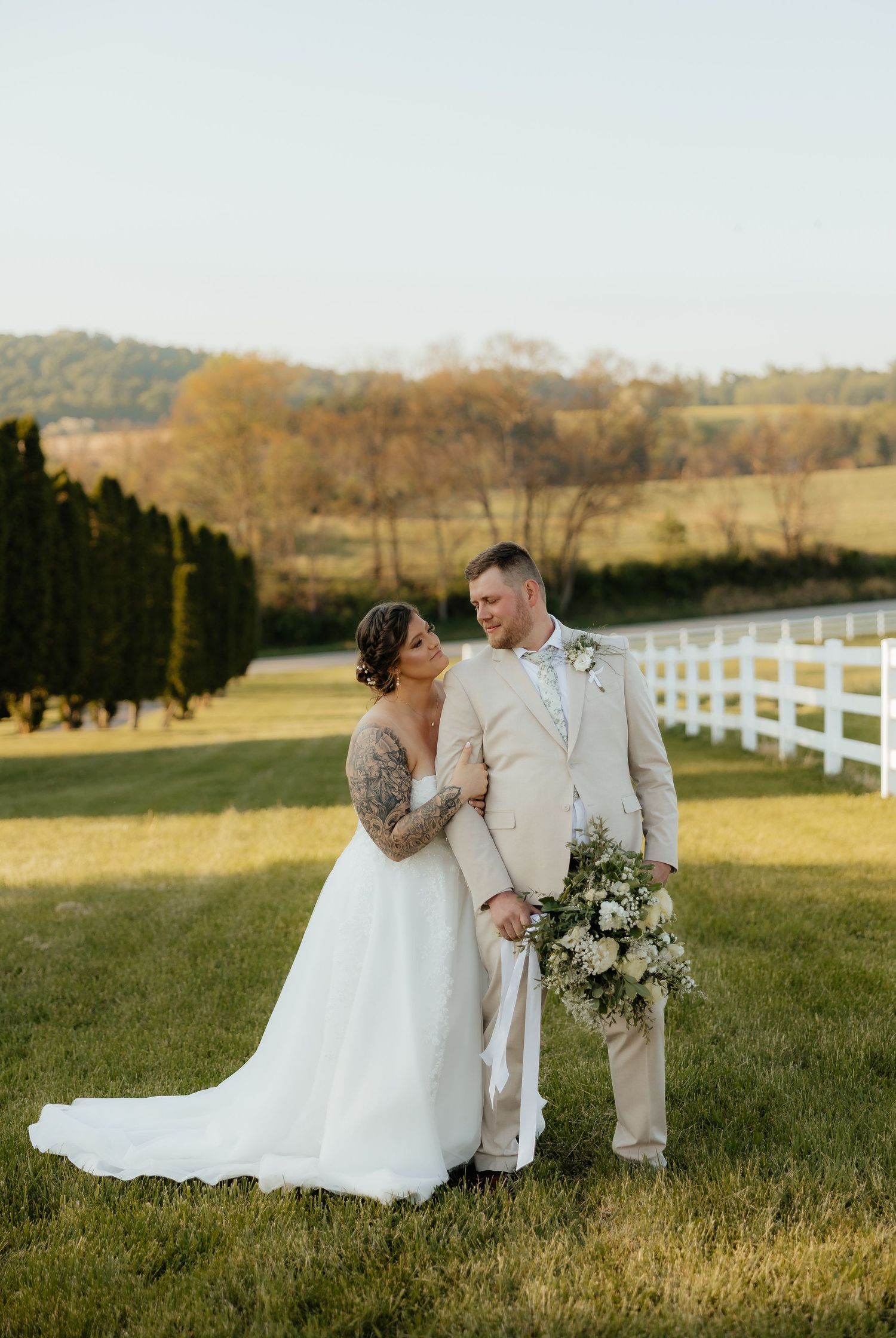 Bride and groom pose on green lawn at golden hour near white fence and rolling hills backdrop.