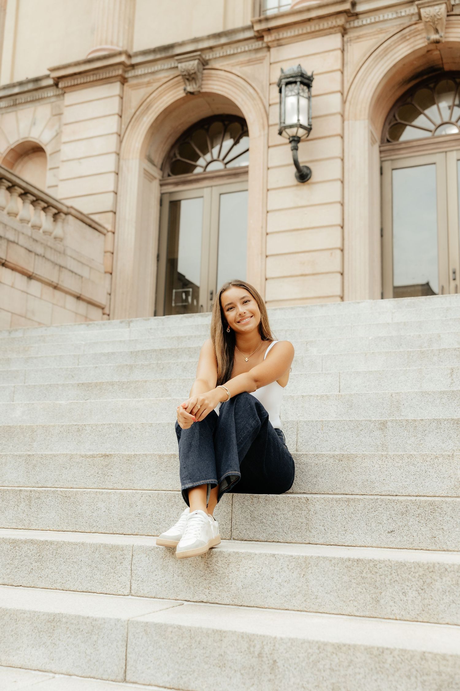 Young woman in casual attire sitting on stone steps of a grand historic building with white sneakers.