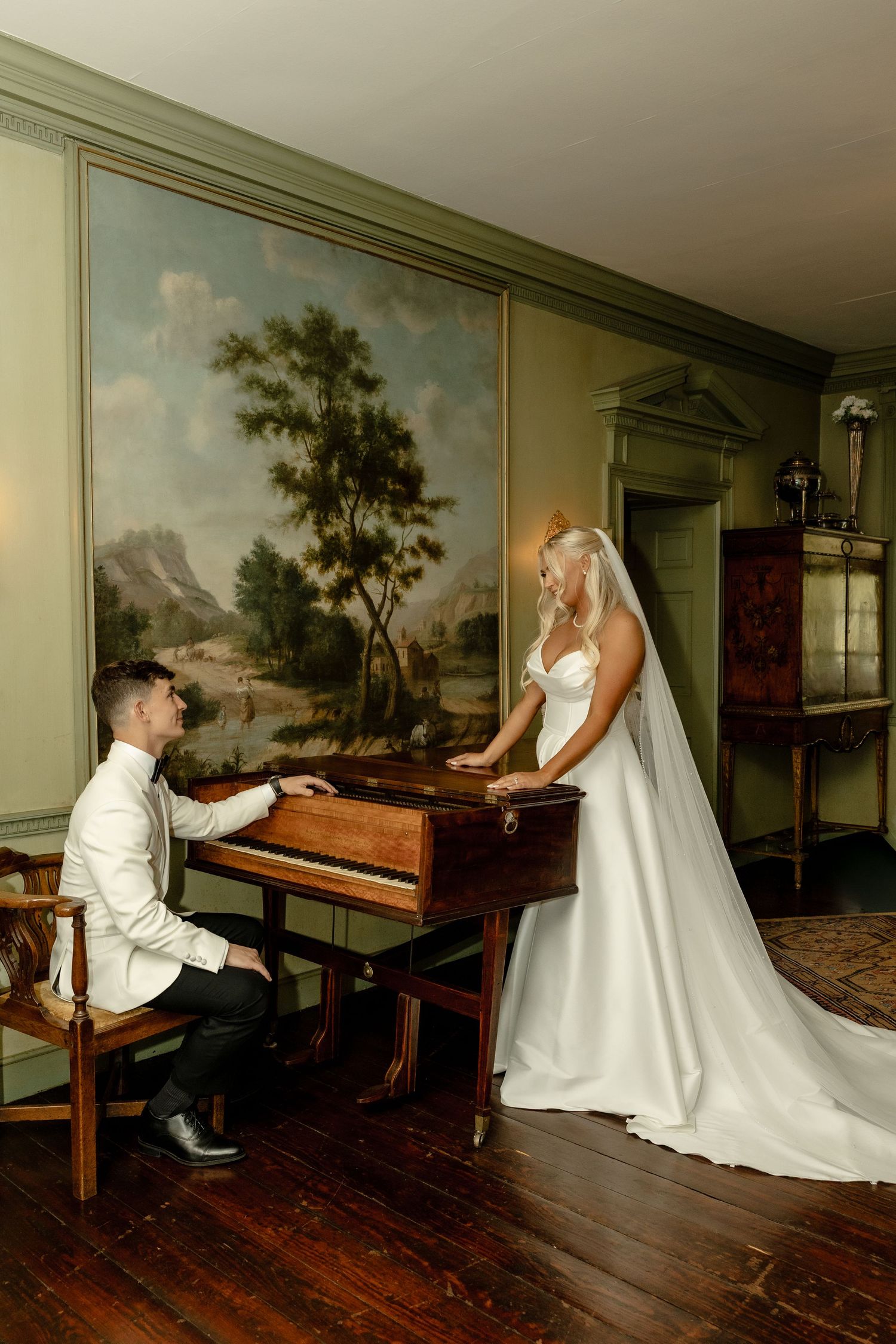 Bride in white gown stands by antique piano as groom plays in elegant room with landscape mural backdrop.