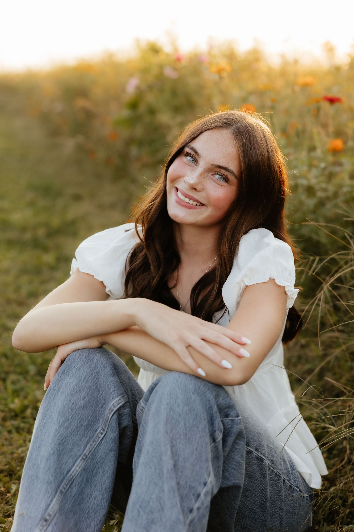 Young woman with brown hair smiling in a flower field, wearing white top and jeans, sitting in golden hour light.