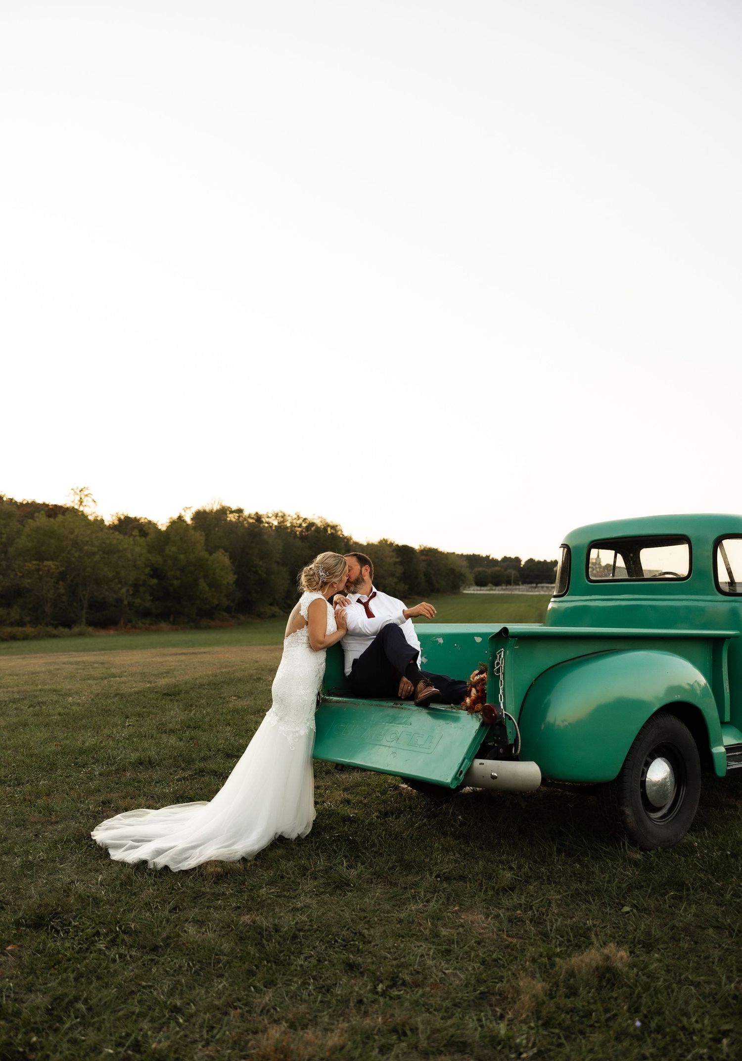 A romantic couple poses with a vintage mint green pickup truck in a rustic field at sunset.