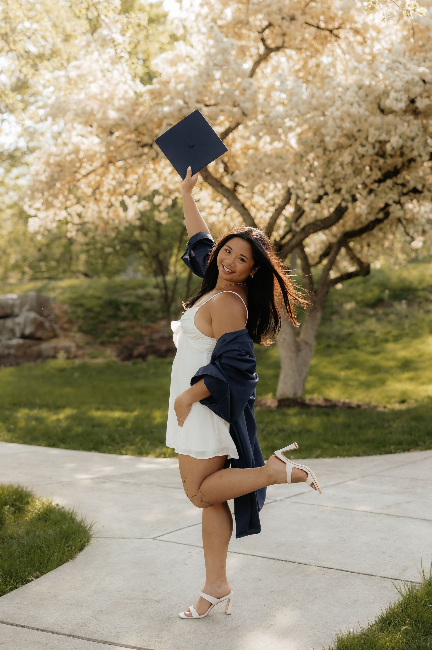 Smiling graduate in white dress holds cap aloft beside blooming cherry trees on a sunny day.