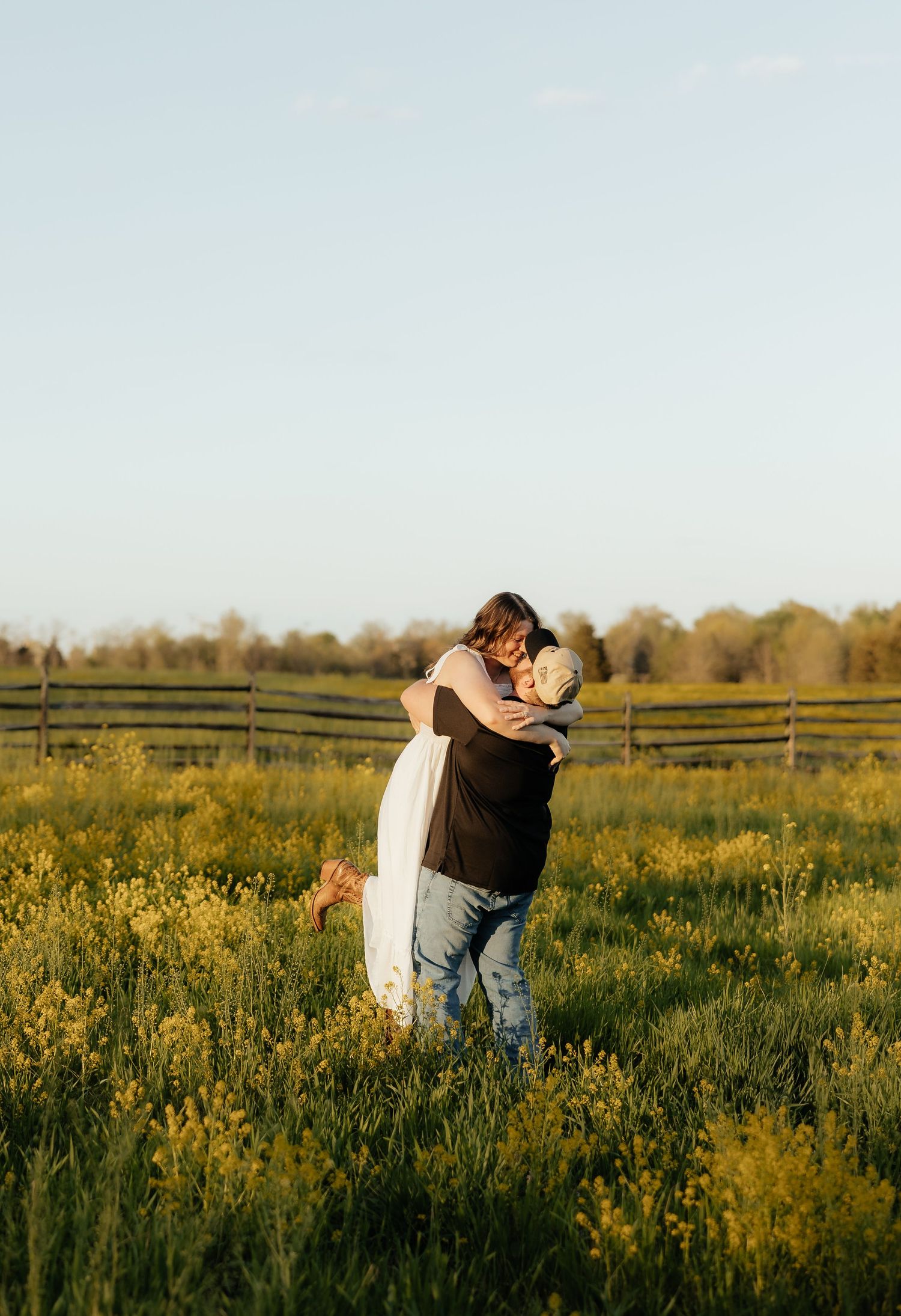 Couple embracing and kissing in a sunlit green field with a wooden fence and trees in the background.