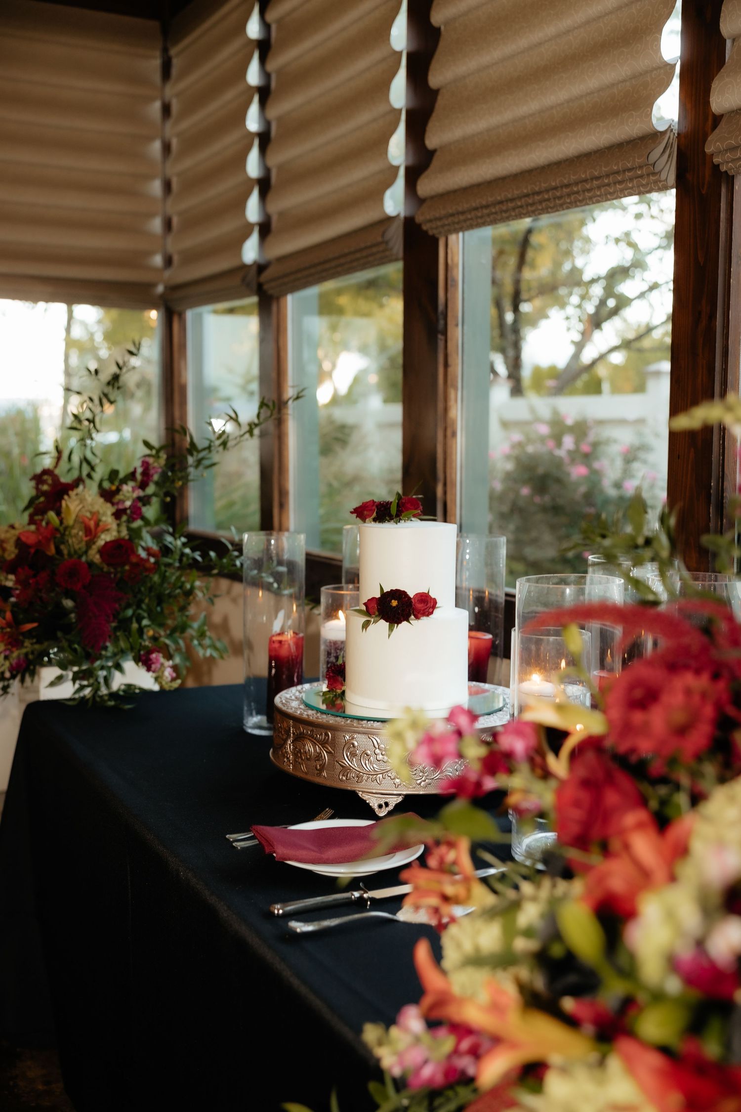 Elegant wedding cake with red floral accents on a gold stand, surrounded by candles and colorful blooms on a dark tablecloth.