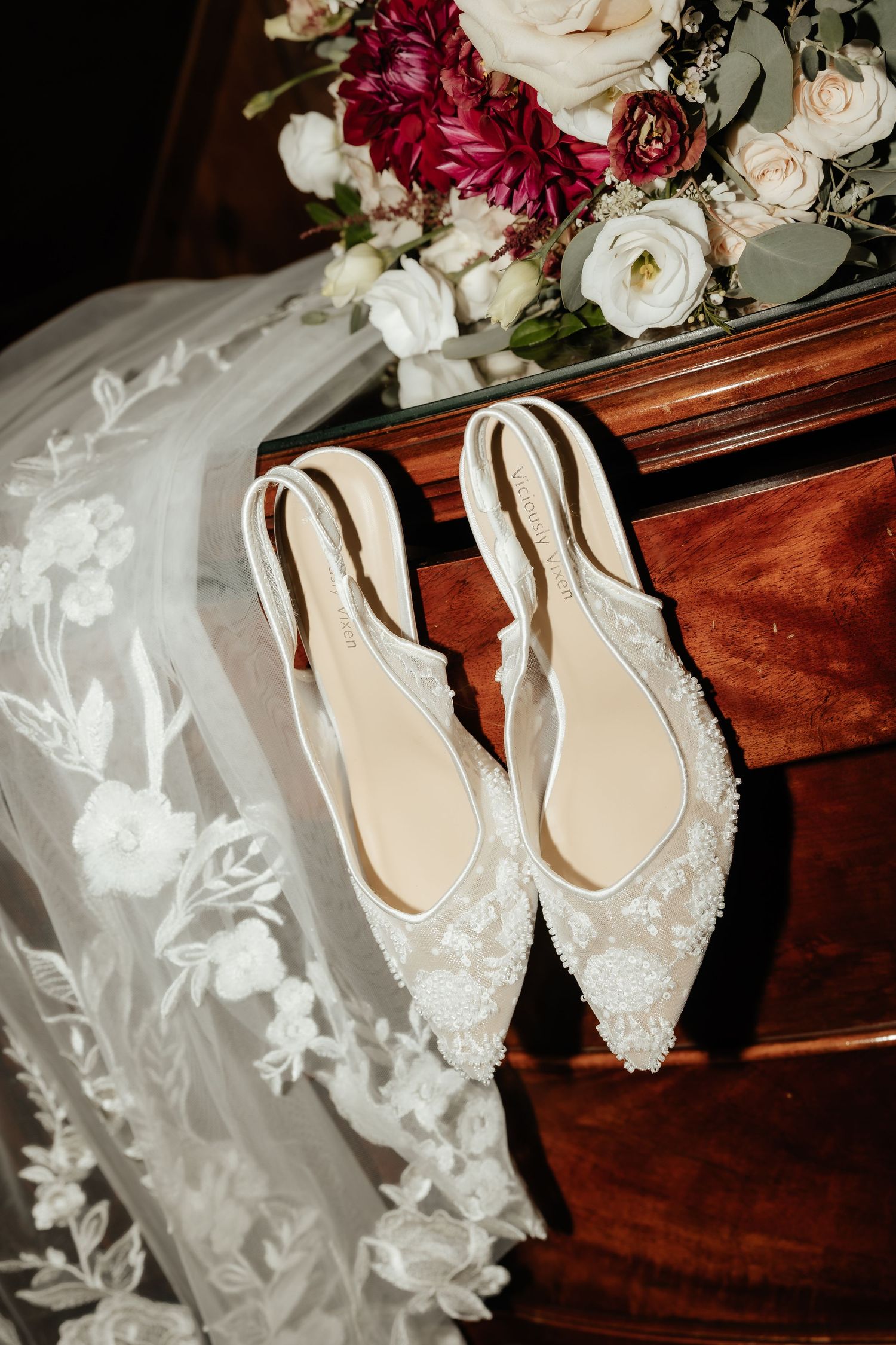 Elegant white lace bridal slingback heels with pearl details on a wooden dresser beside a floral bouquet and embroidered veil.