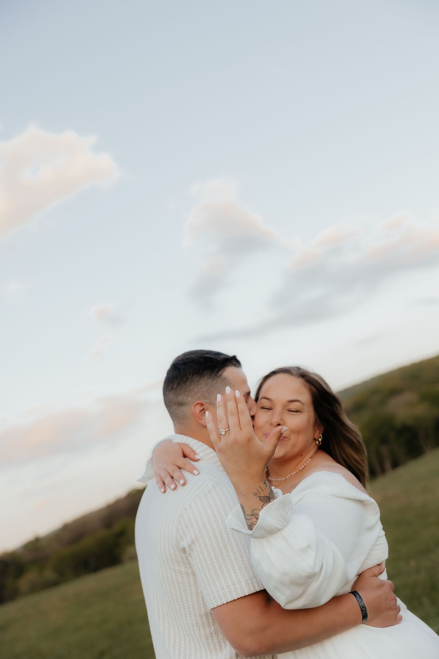 A couple shares a romantic kiss outdoors, woman showing engagement ring, surrounded by green hills and soft cloudy sky.