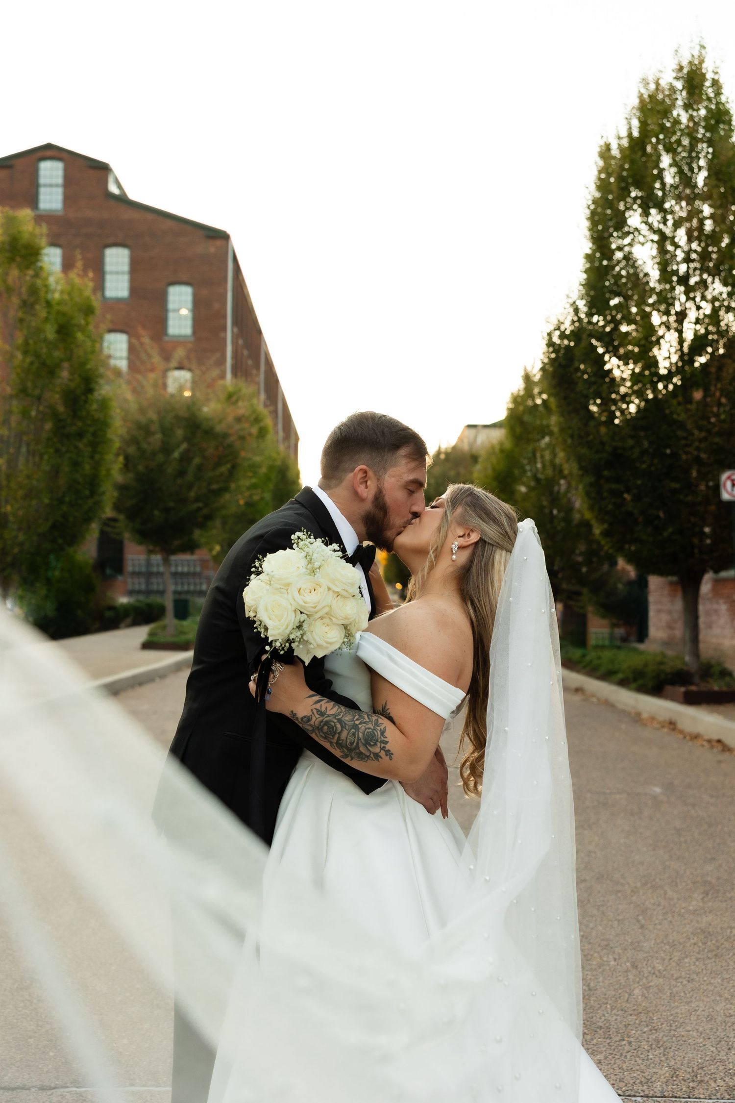 A romantic wedding couple shares a kiss outdoors with a rustic brick building and trees in the background.