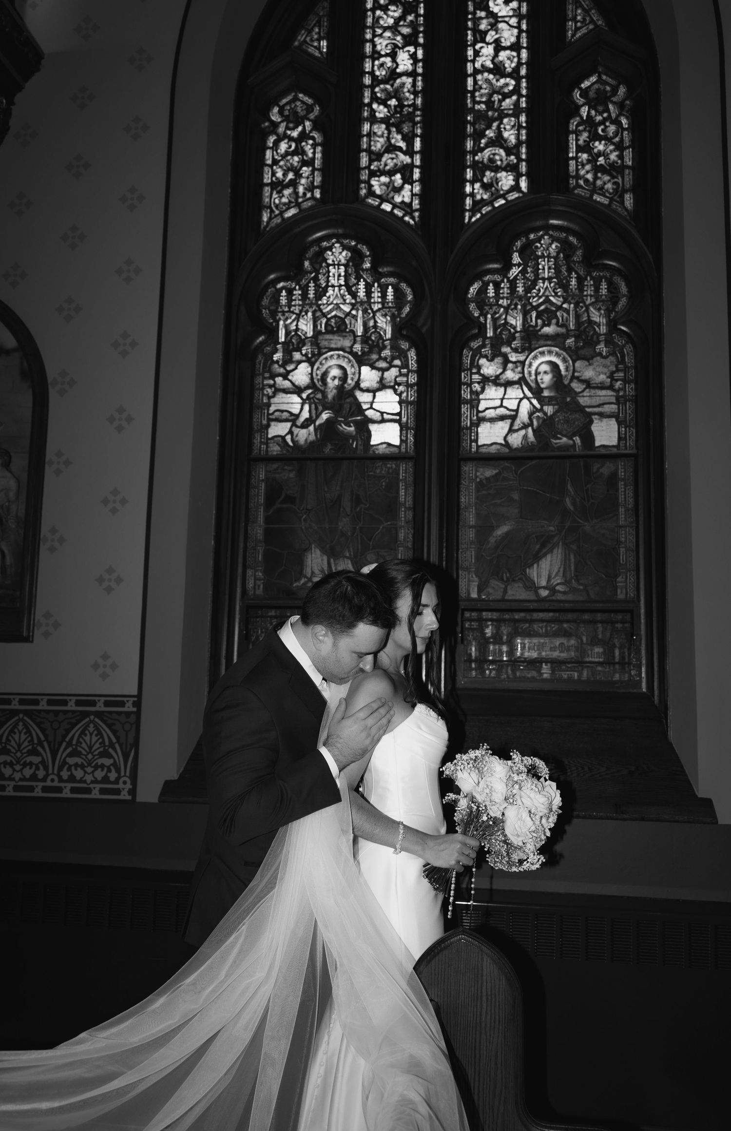 Black and white photo of a bride and groom sharing a kiss in front of stunning stained glass windows in a church.