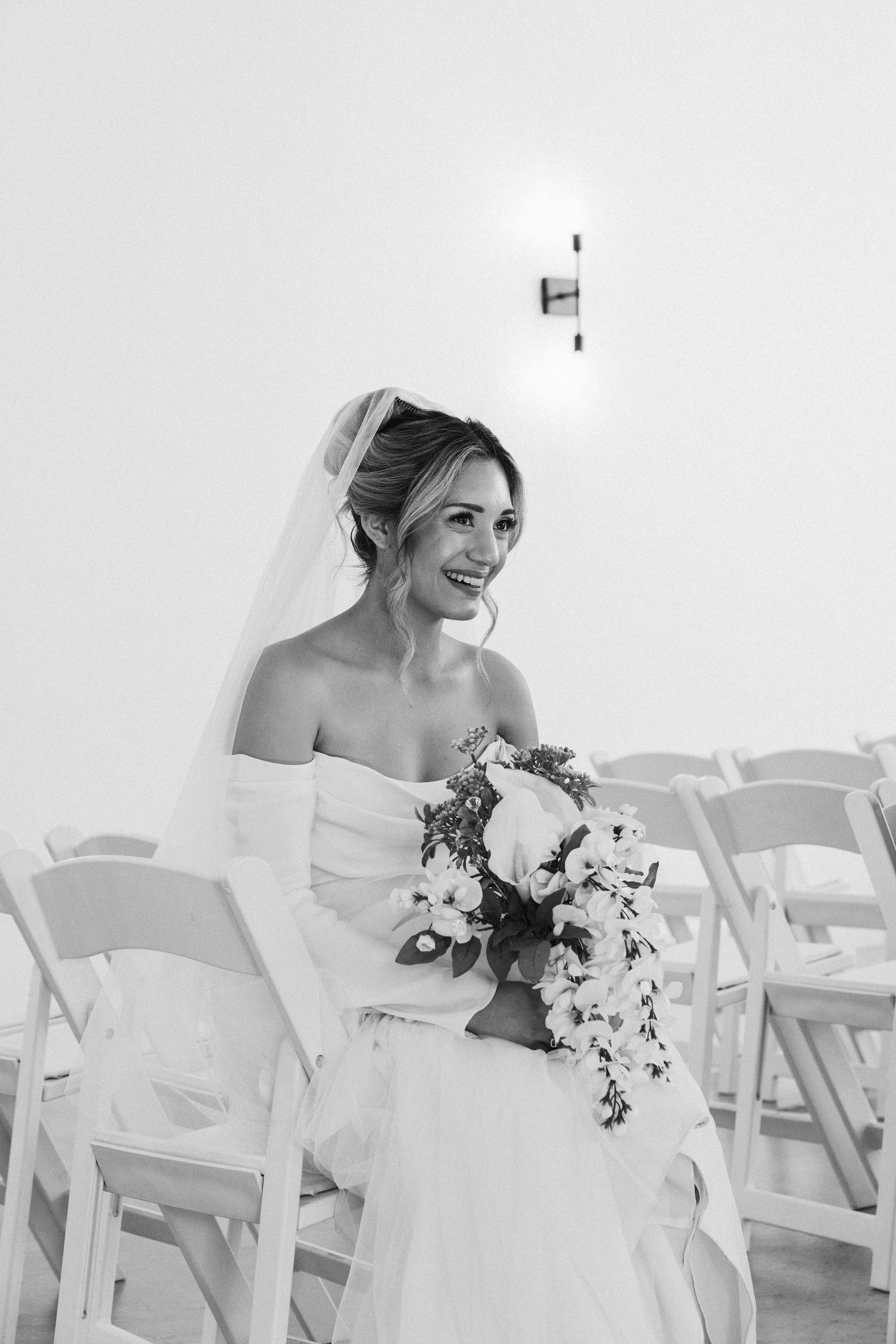 Black and white wedding portrait of a smiling bride holding a cascading bouquet while seated in ceremony chairs.