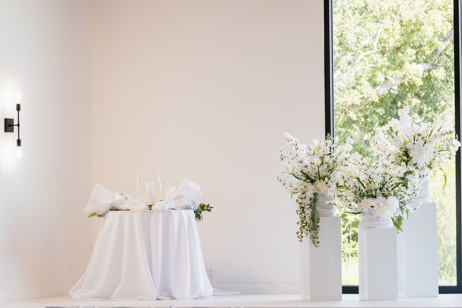 Elegant white wedding reception setup featuring a sweetheart table and white floral arrangements on pedestals near a large window.