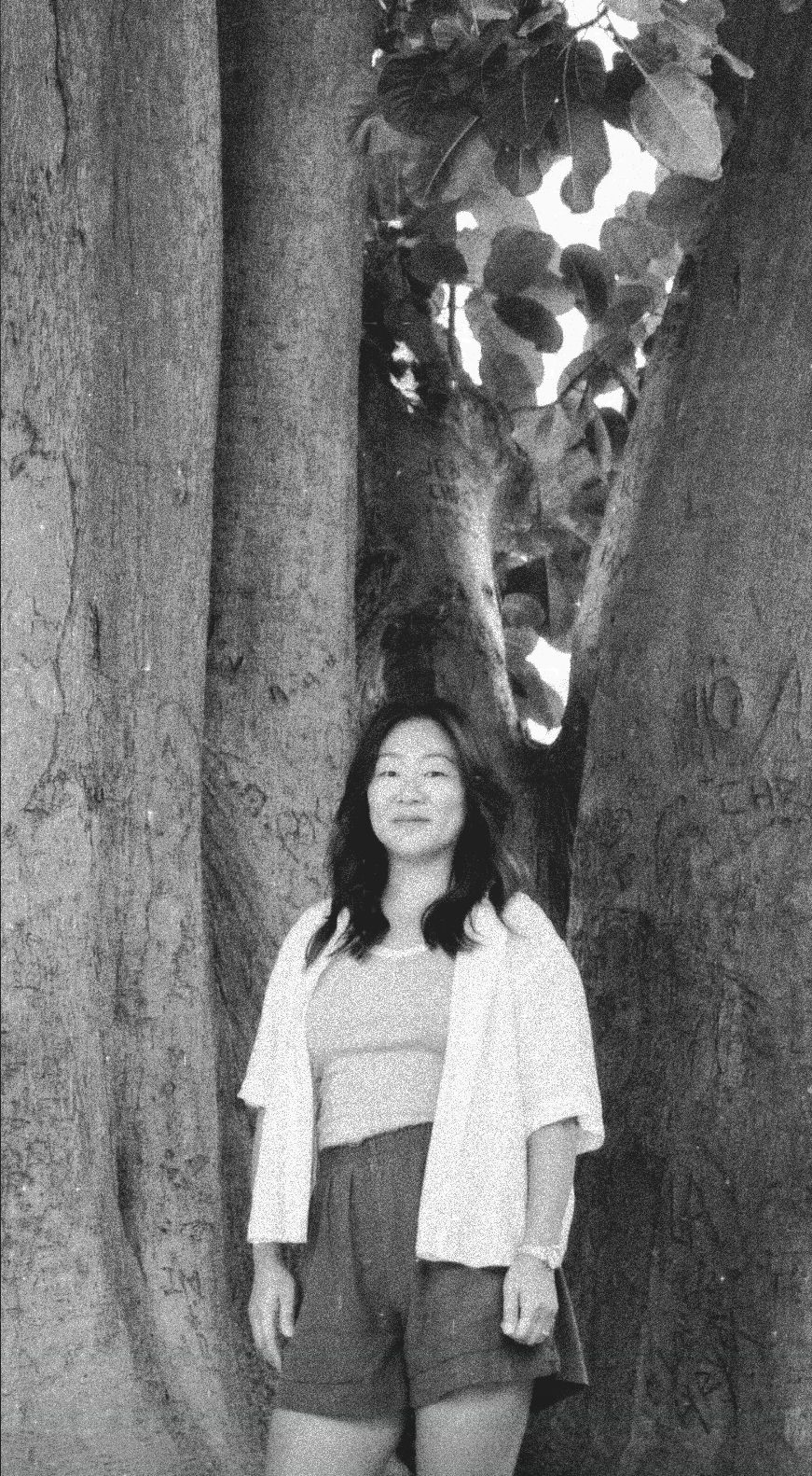 Black and white outdoor portrait of a smiling woman standing framed between large, rustic tree trunks with leaves above.