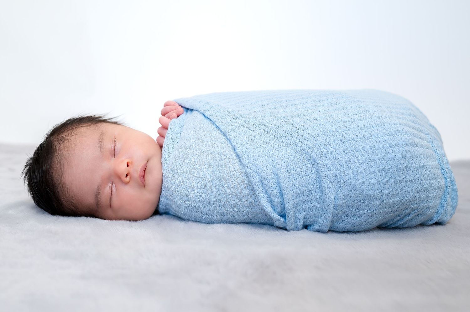 A peacefully sleeping newborn swaddled in soft blue striped fabric lies on a gray blanket.