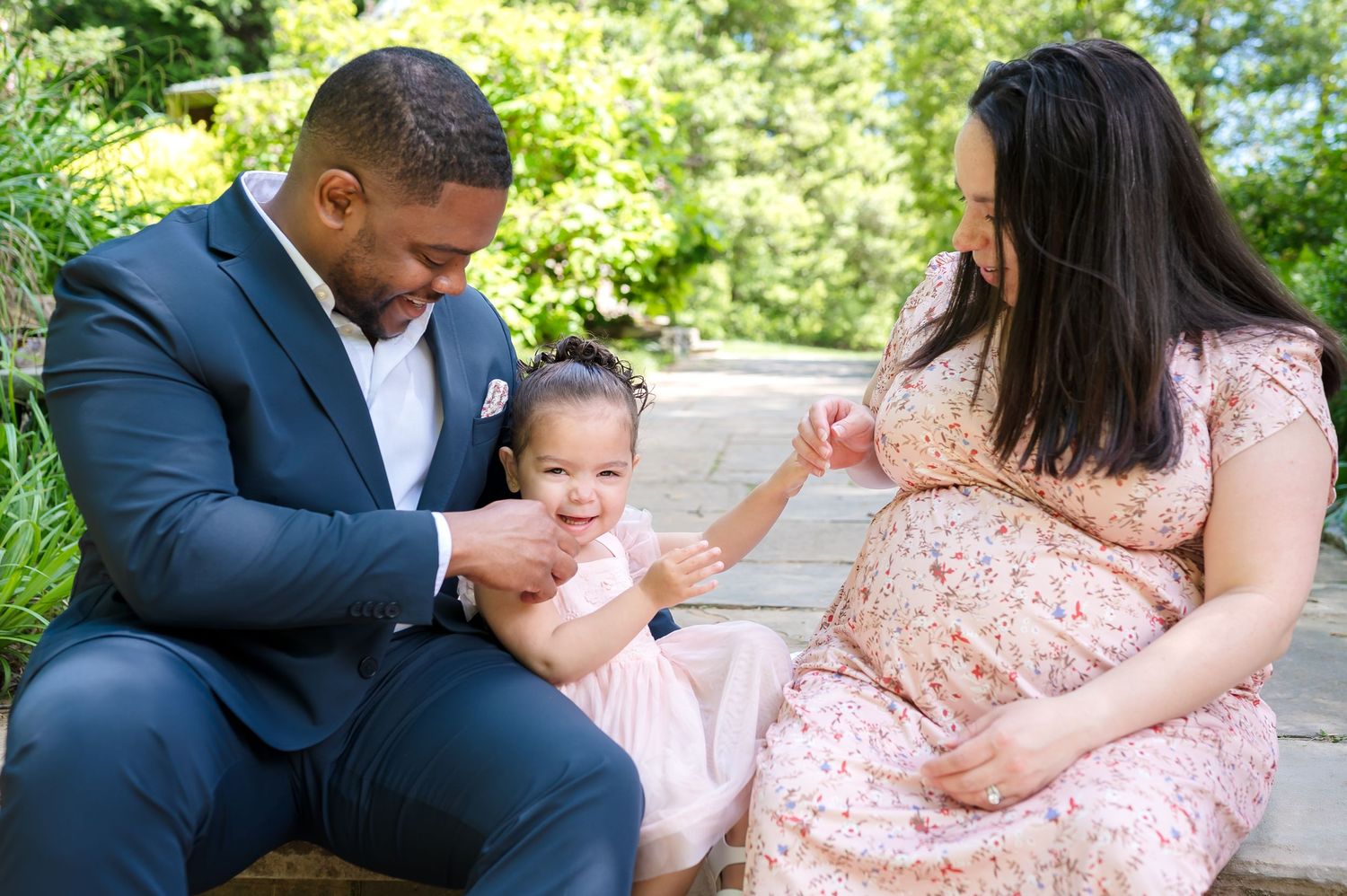 Family wearing coordinated navy and pink outfits poses together outdoors in a sunny garden setting.