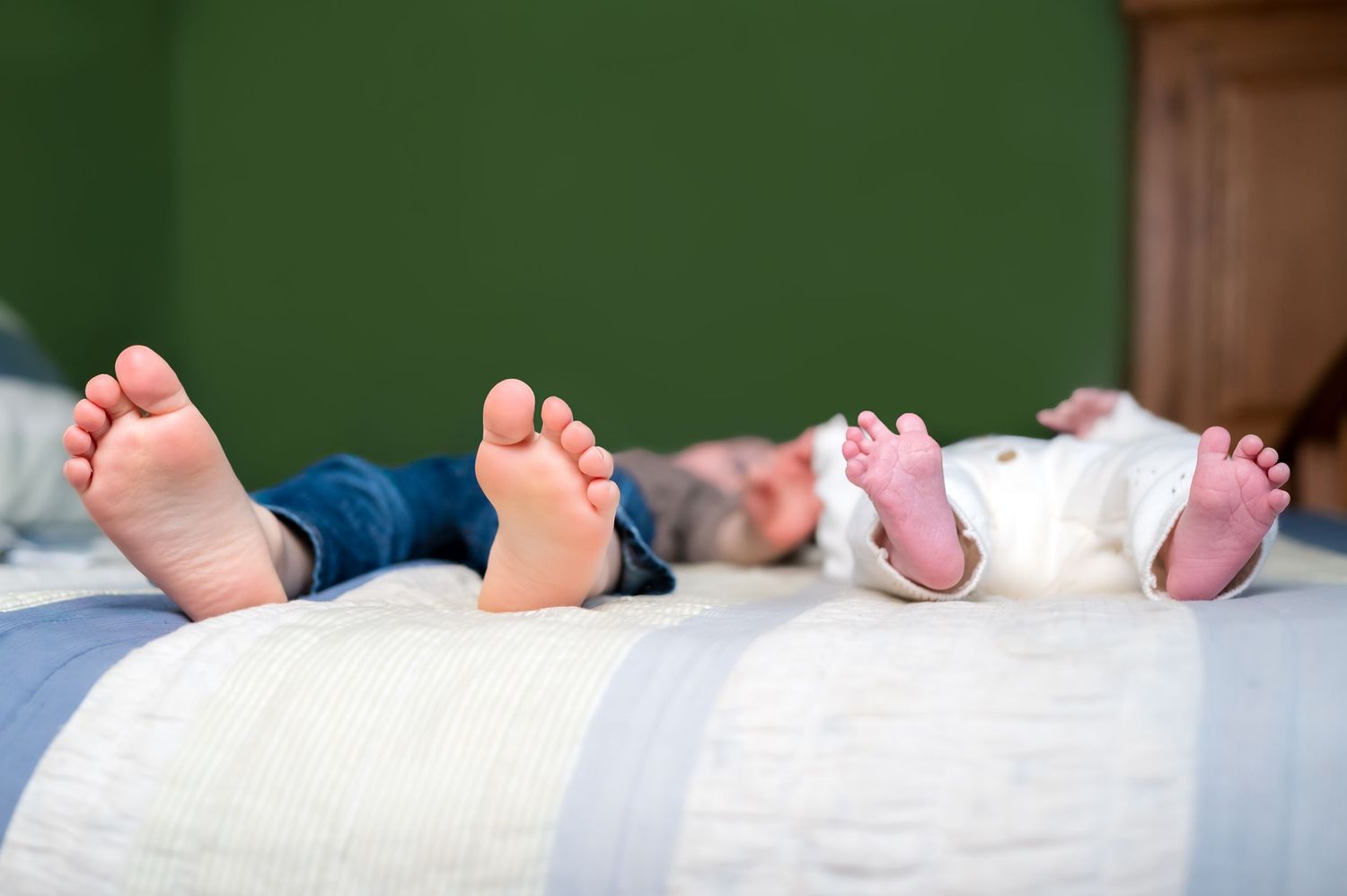 Two children lying on a bed with bare feet up, a toddler and newborn baby side by side on white bedding.