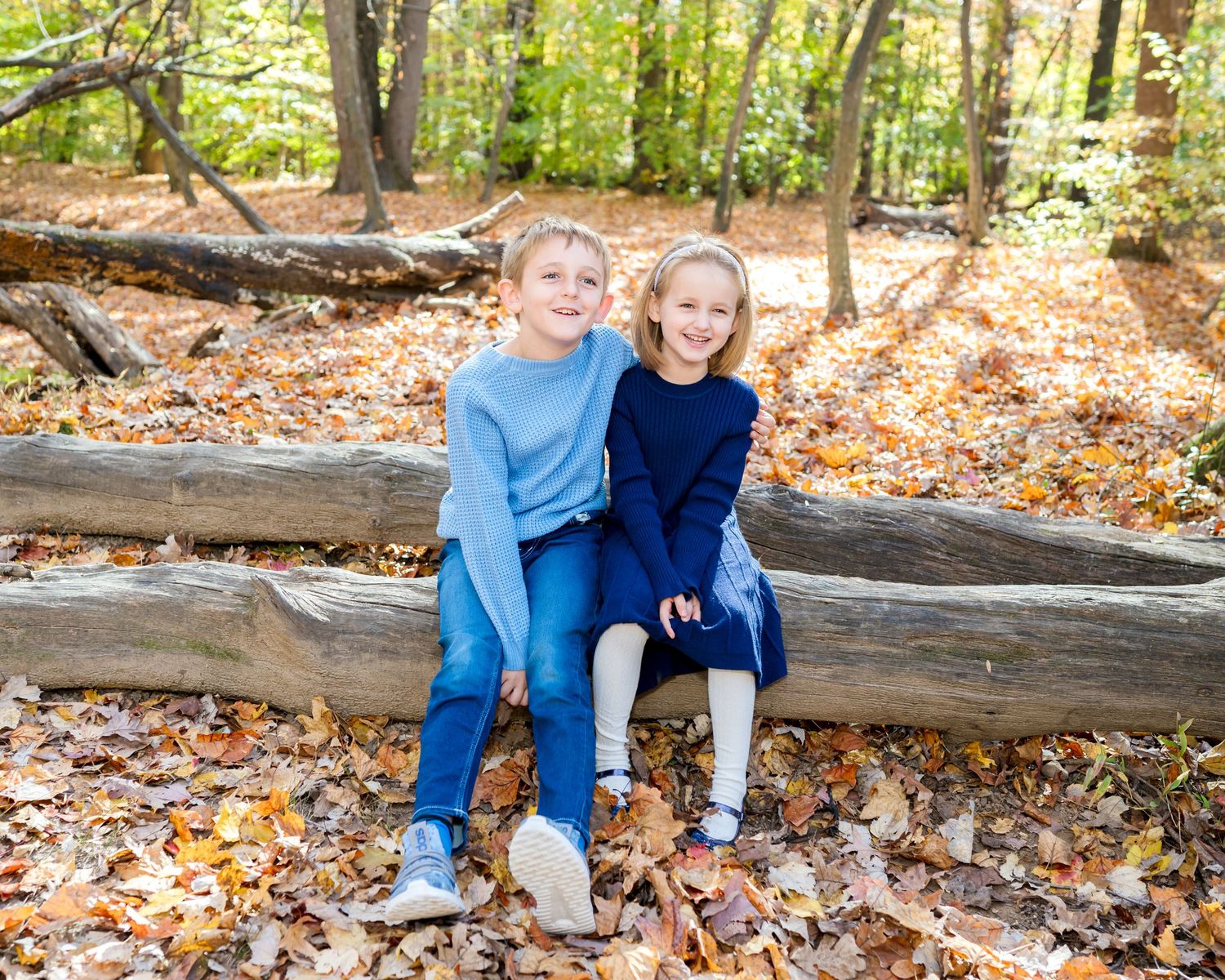 Two siblings sit together on a fallen log in a forest of colorful autumn leaves during an outdoor photoshoot.