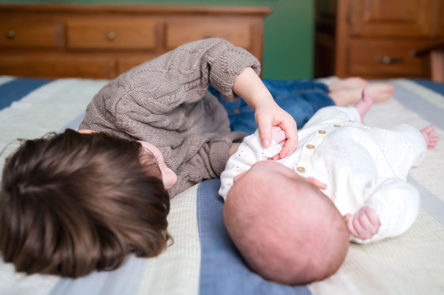 Toddler girl leaning over newborn baby sibling lying on a bed with blue striped blanket.