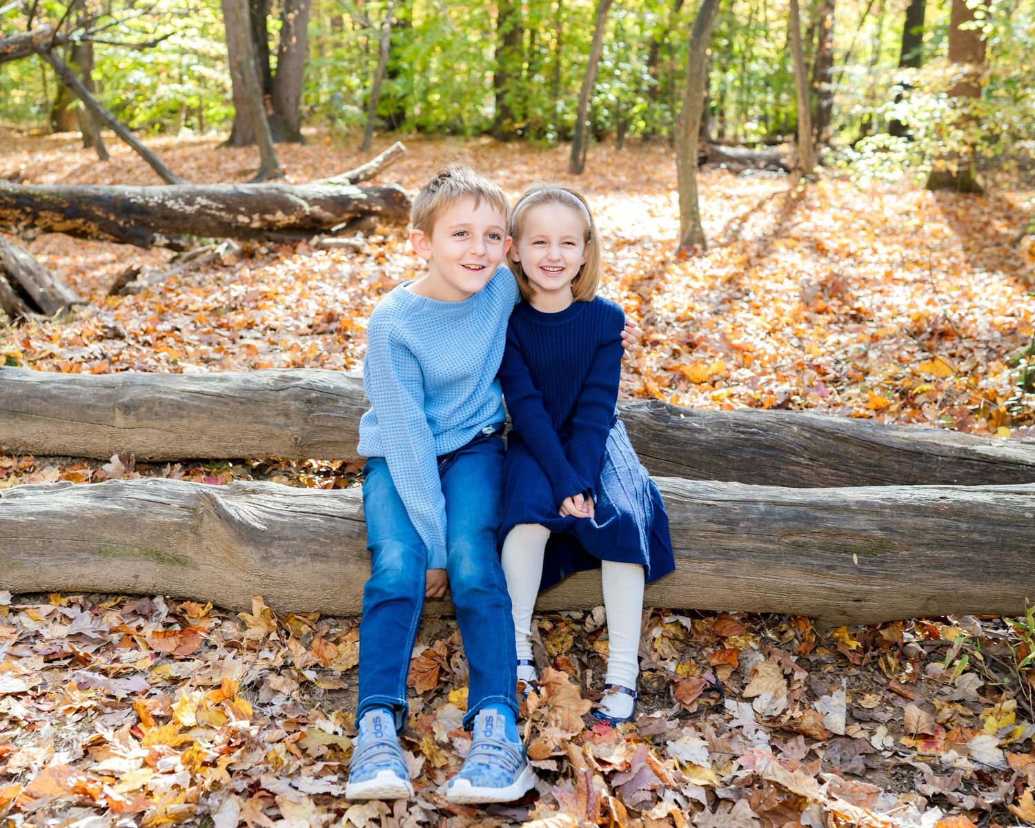Two children in blue sit together on fallen logs surrounded by autumn leaves in a wooded forest area.