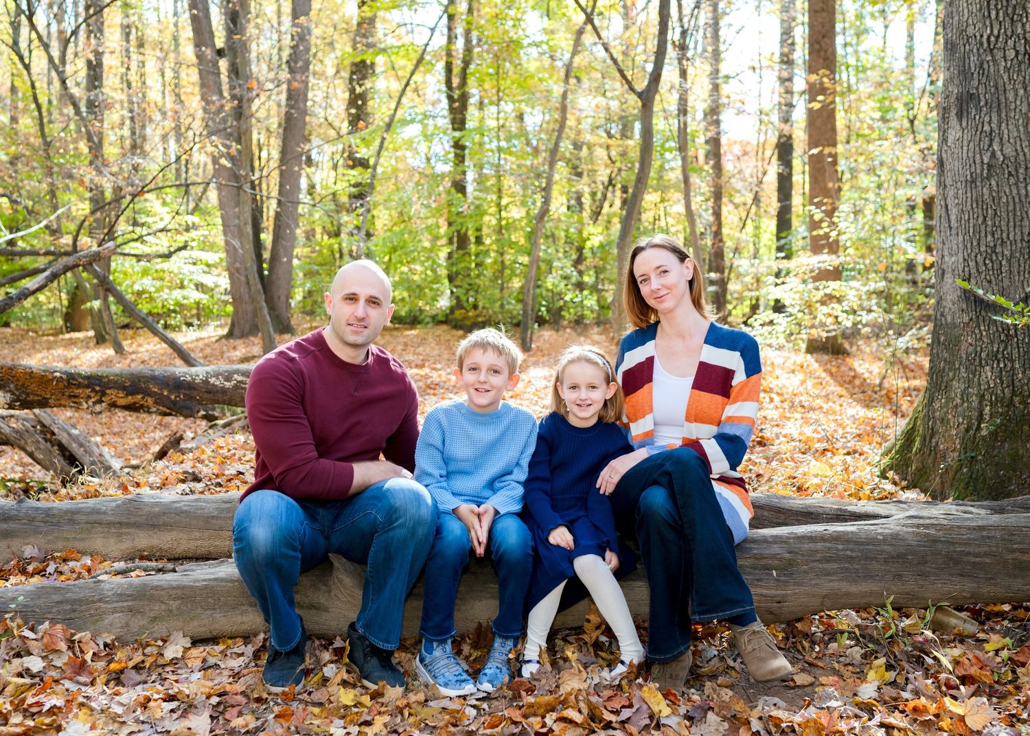 Family group sitting together on fallen logs in an autumn forest with colorful leaves scattered on the ground.