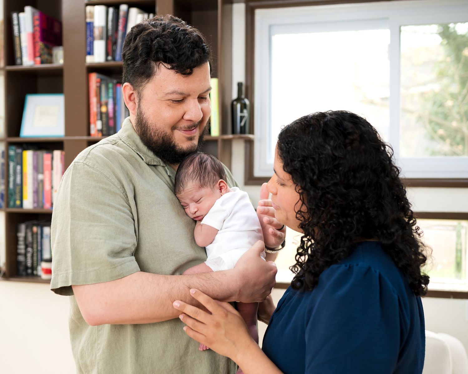 A couple tenderly holds their newborn baby together in a cozy home library setting.