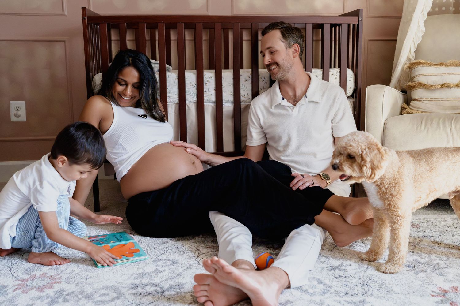 Pregnant woman sits on nursery floor with toddler and husband, smiling near crib with a fluffy dog beside them, in Odenton, MD