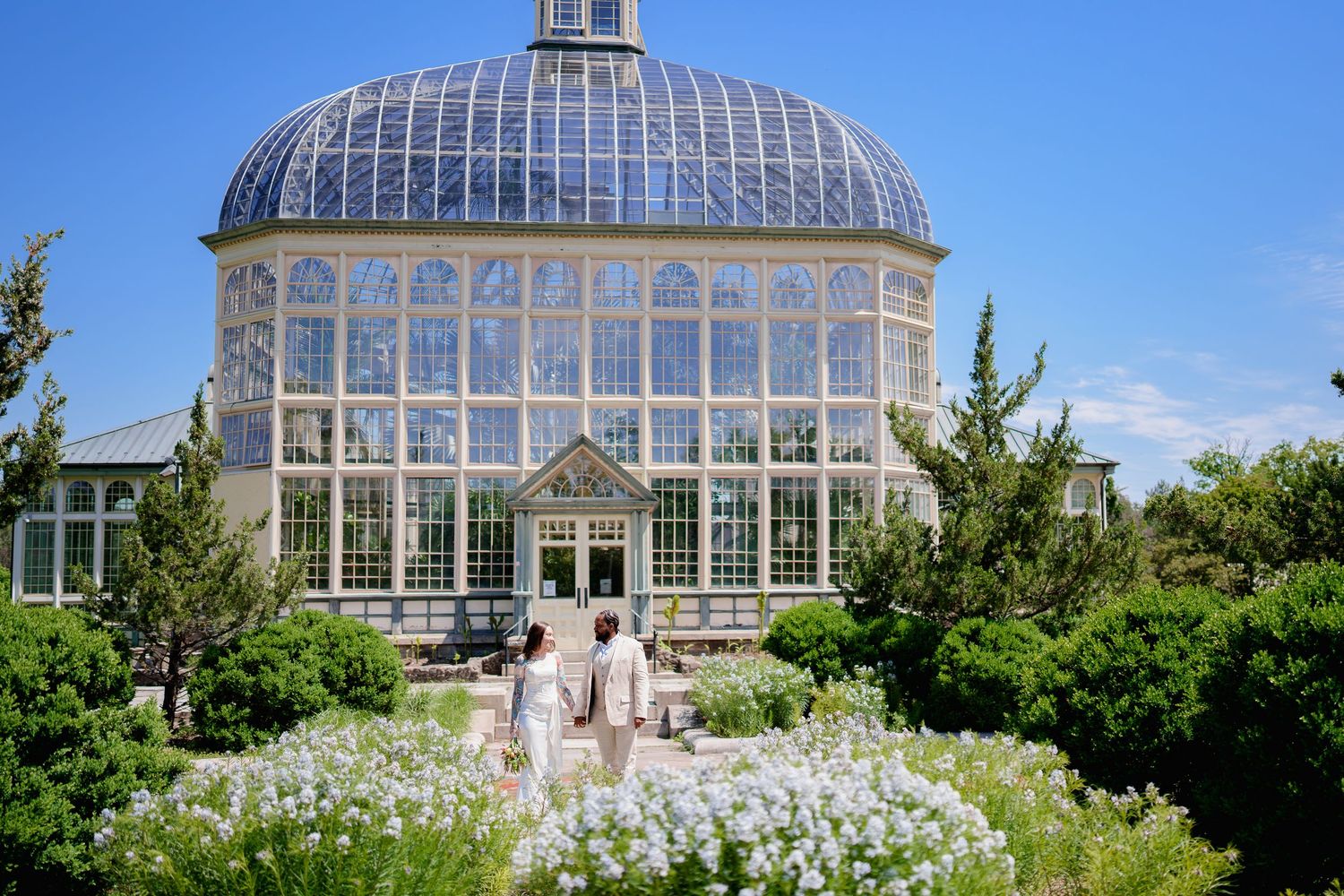 Married couple walks toward a grand Victorian glass greenhouse surrounded by lush white flowering gardens under a bright blue sky.