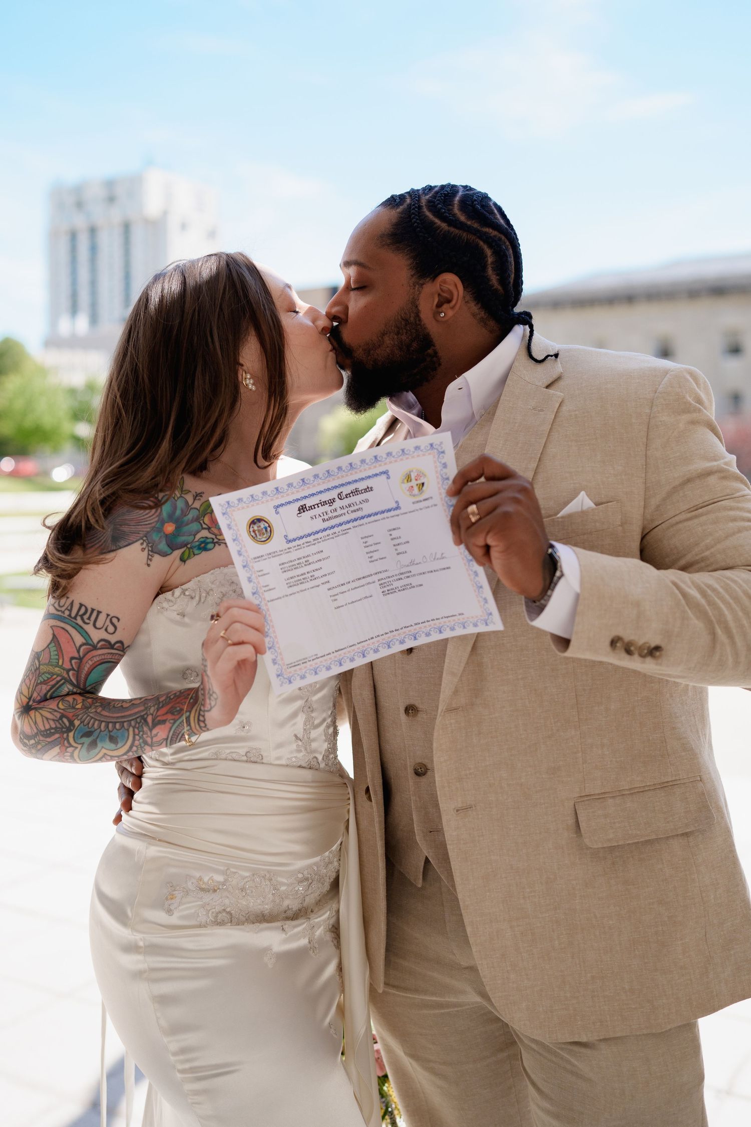 Newlywed interracial couple kissing while holding marriage certificate outdoors, bride with tattoos in white gown, groom in beige suit, MD