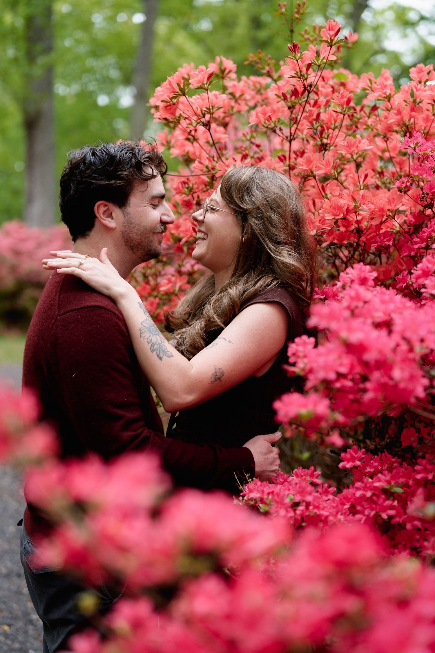 Couple embracing and smiling among vibrant pink azalea bushes in a lush green garden setting, during engagement session in Brookeville, MD