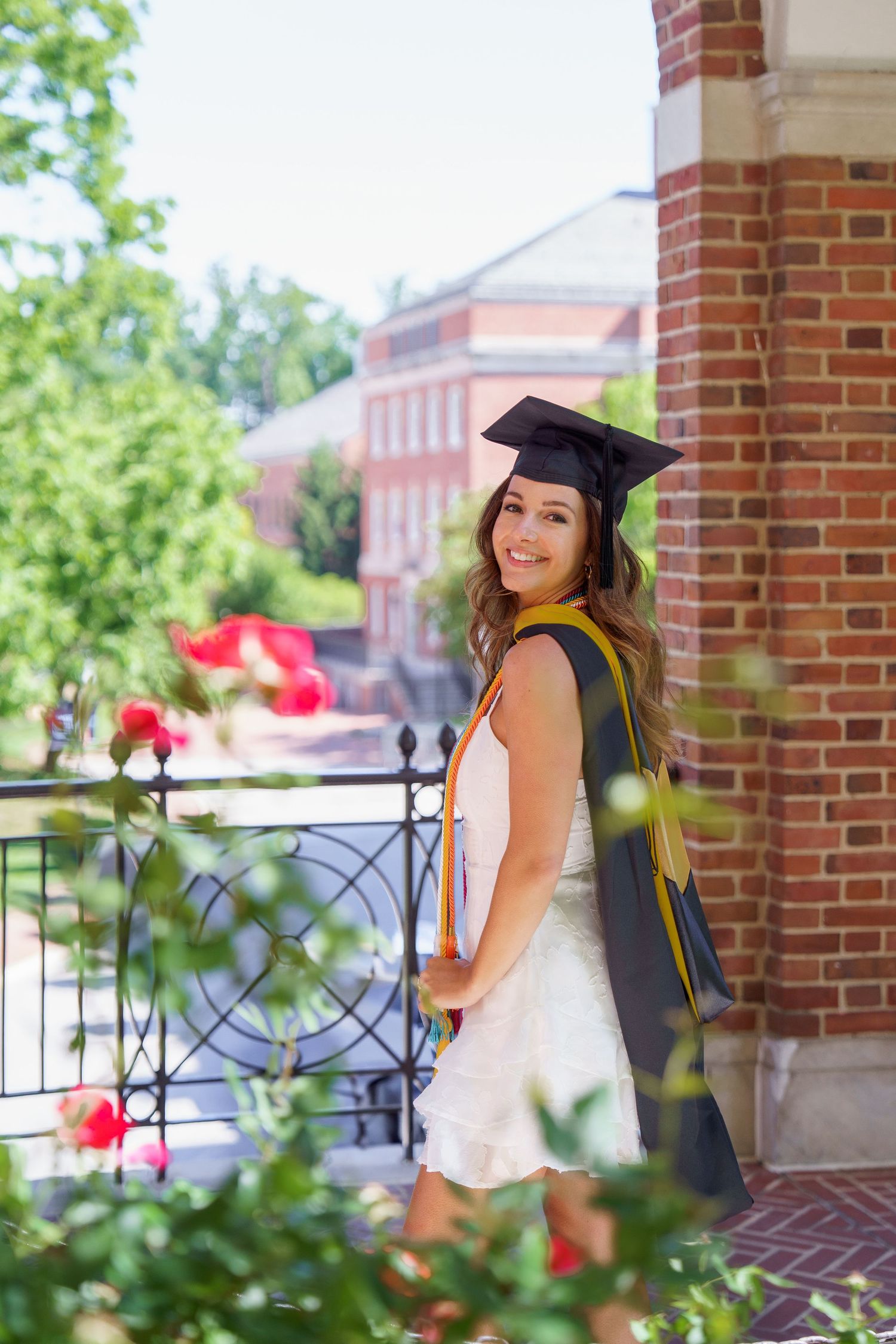 Graduate poses in white dress and cap with red flowers and brick columns on a sunny college campus.