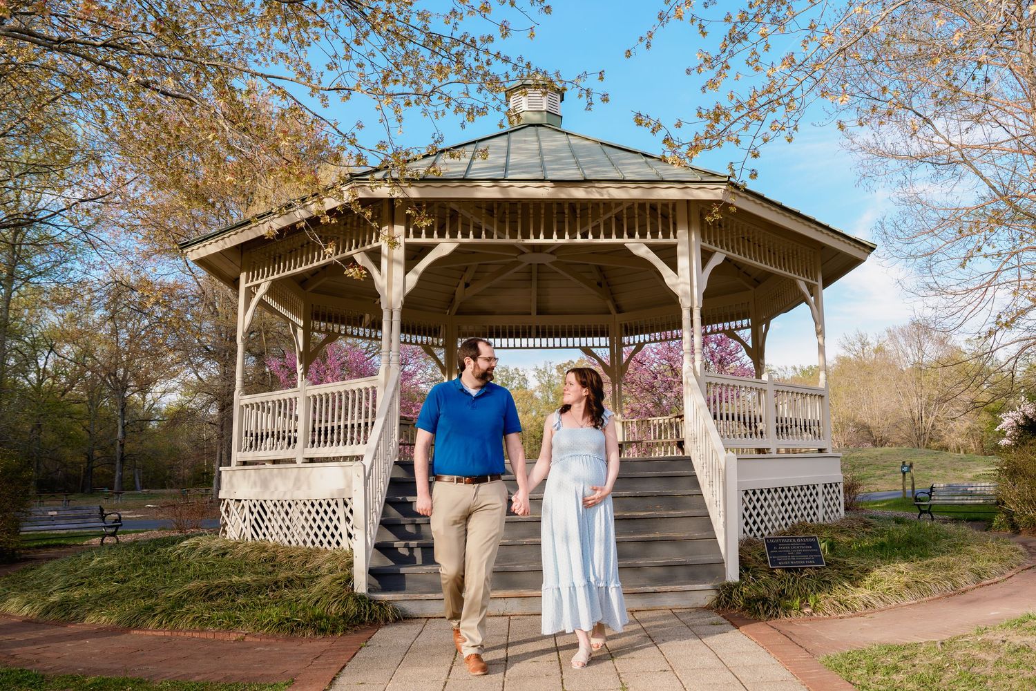 Pregnant couple walking hand-in-hand near a white gazebo surrounded by spring trees in a park, during maternity session in Annapolis, MD