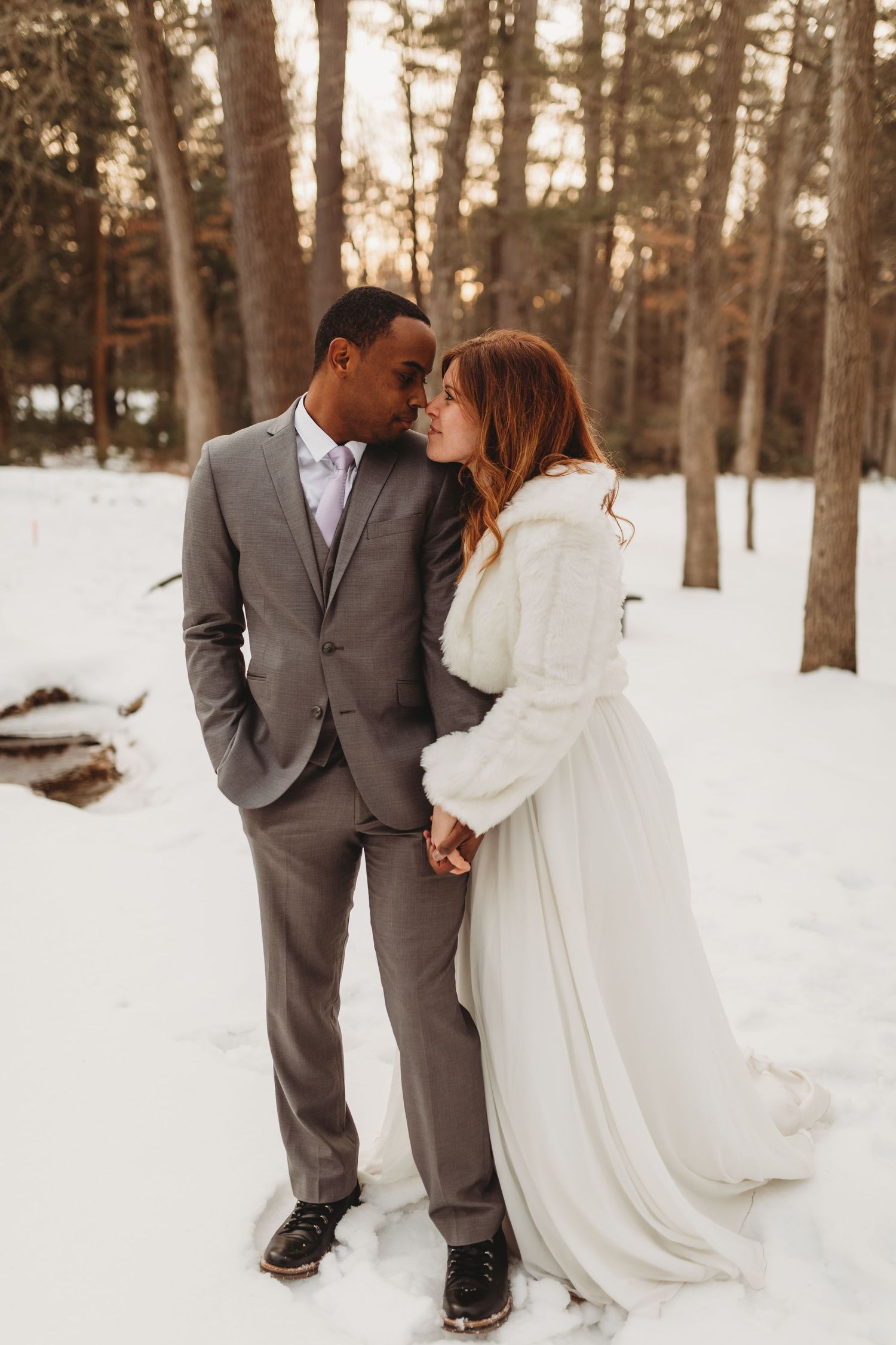 Interracial couple in winter wedding attire share a romantic moment in a snowy forest setting