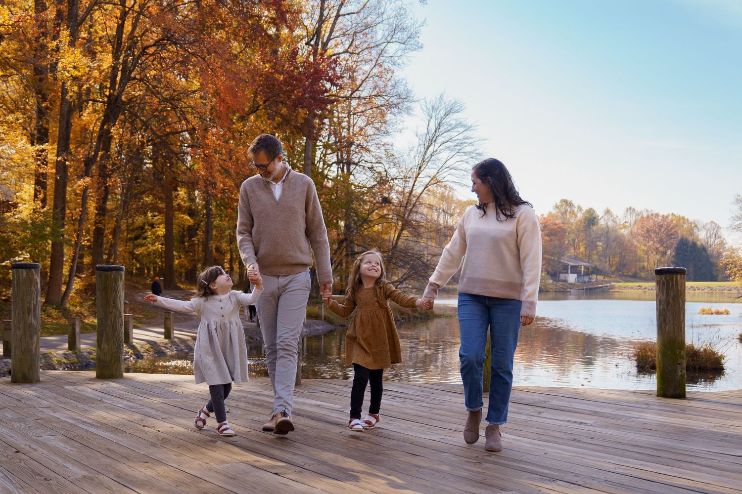 Family of four holding hands walking on wooden dock surrounded by autumn foliage near a lake on a sunny day, in Columbia, MD