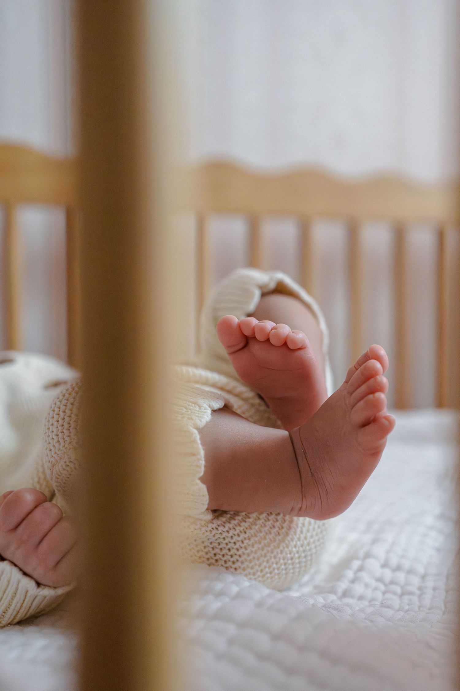 Close-up of newborn baby feet and tiny toes in a white knit outfit, lying in a wooden crib, in Fairfax, VA