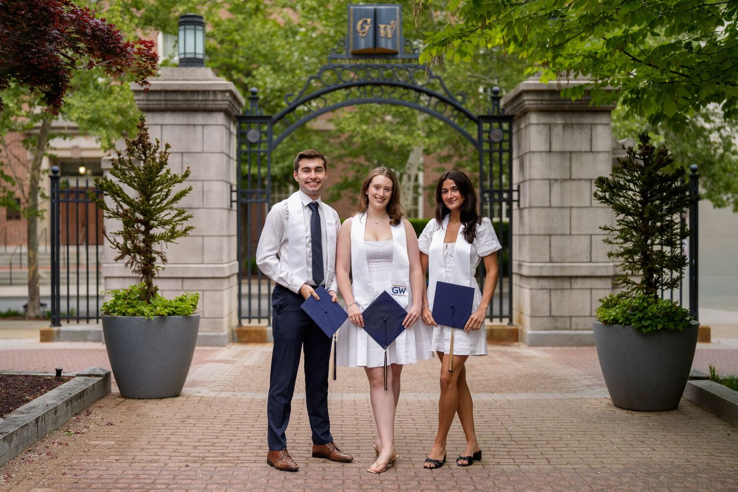 Three graduates in formal attire pose proudly before an ornate gate on a brick campus pathway, George Washington University, in DC