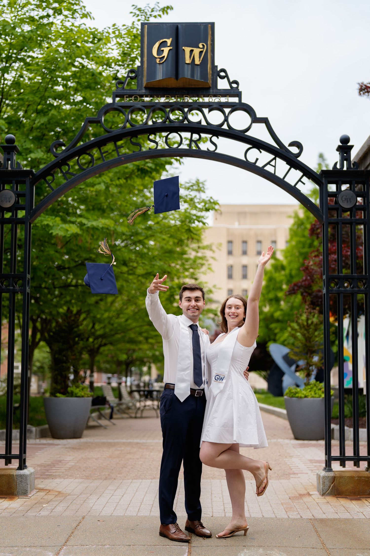 Two GWU graduates celebrate under the Professors Gate, tossing caps in the air on a tree-lined campus path, in Washington DC