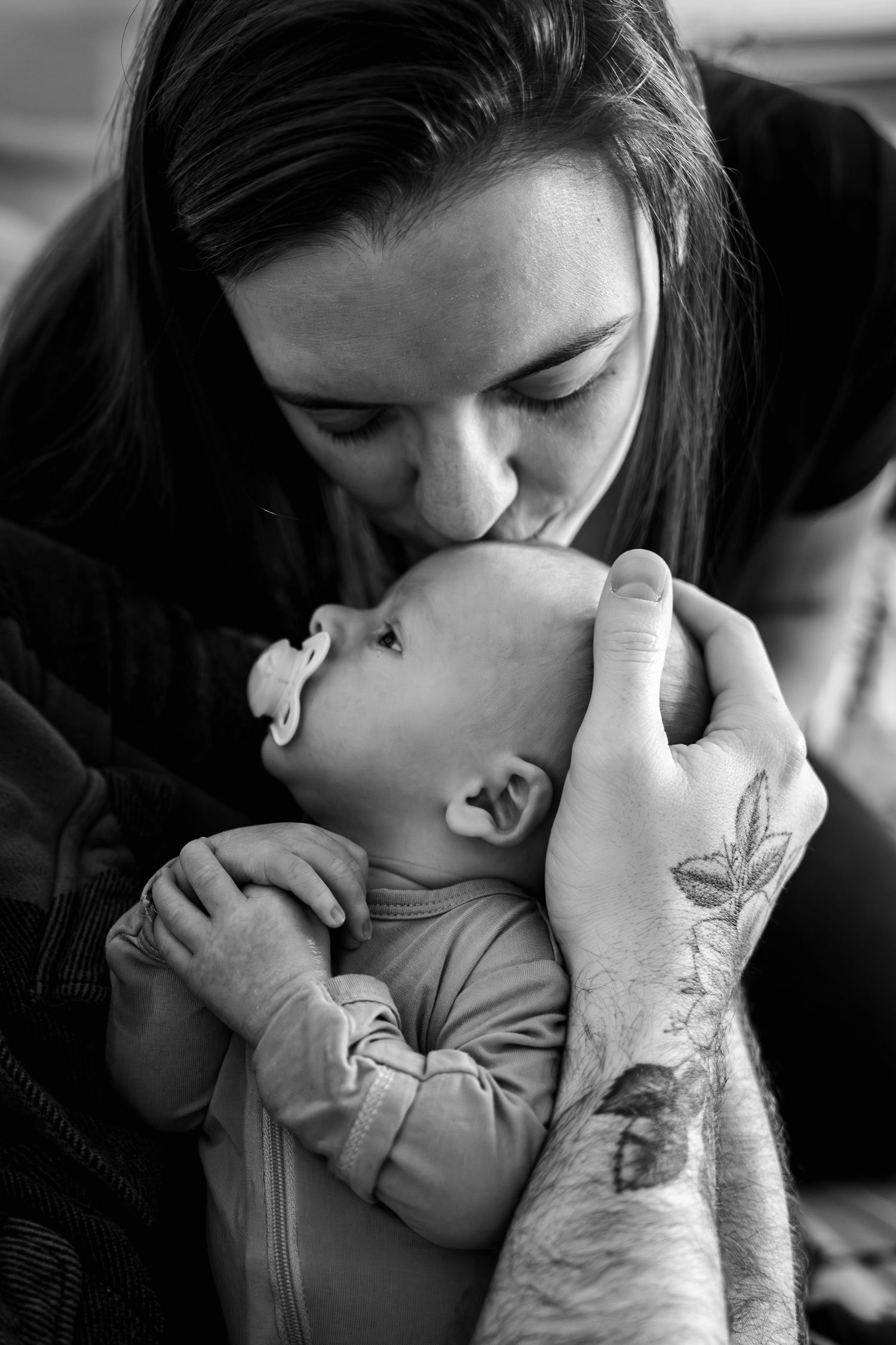 Black and white photo of a mother tenderly kissing her newborn baby with a pacifier, held by tattooed hands, in Columbia, MD